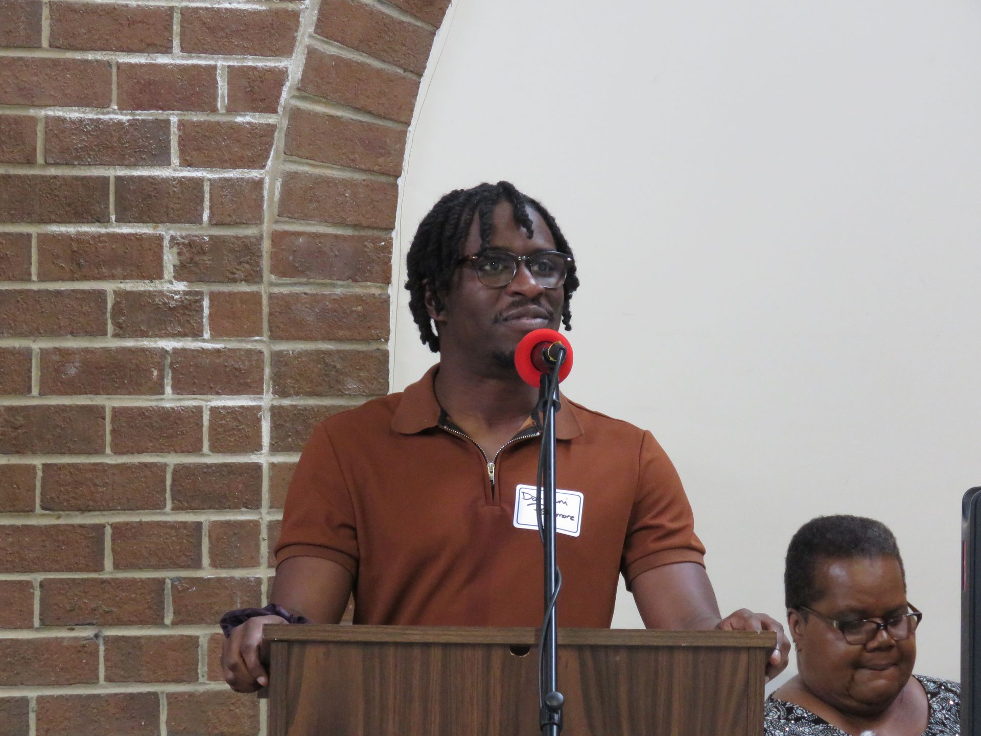 Man speaking at a podium with a microphone, brick wall background, another person partially visible.