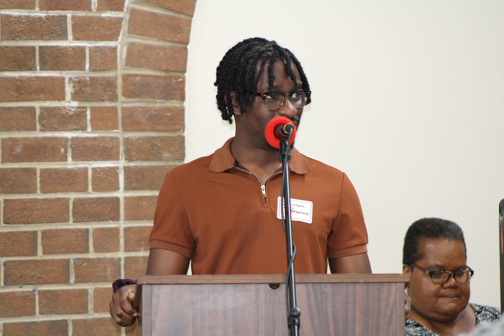 Person speaking at a podium with microphone. Brick wall background.
