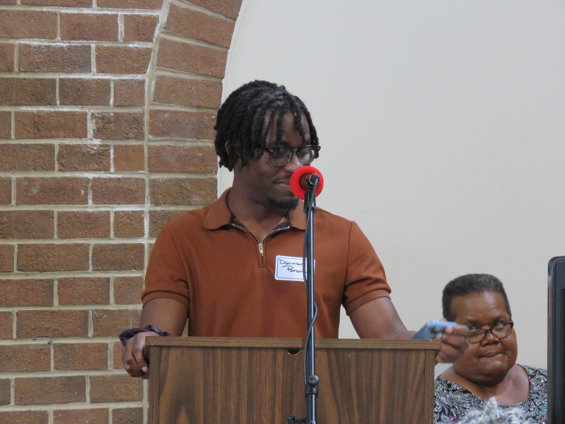 Man speaking at a podium with microphone, brick wall, and woman in background.