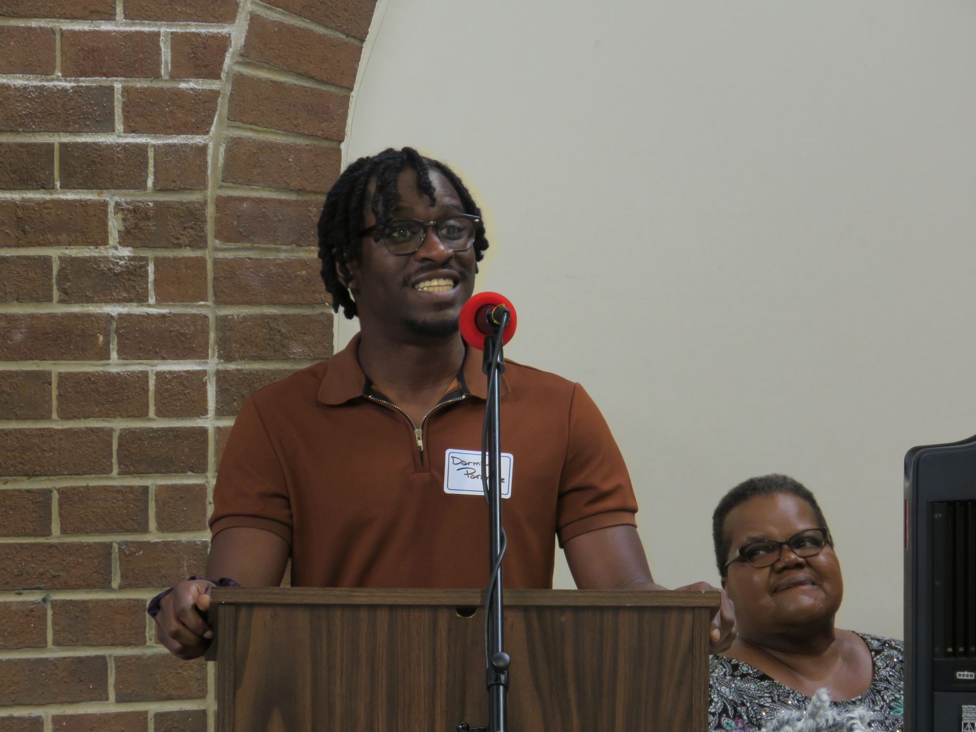 Man speaking at podium, brown shirt, dreadlocks, smiling, brick wall background. Woman seated to the side.