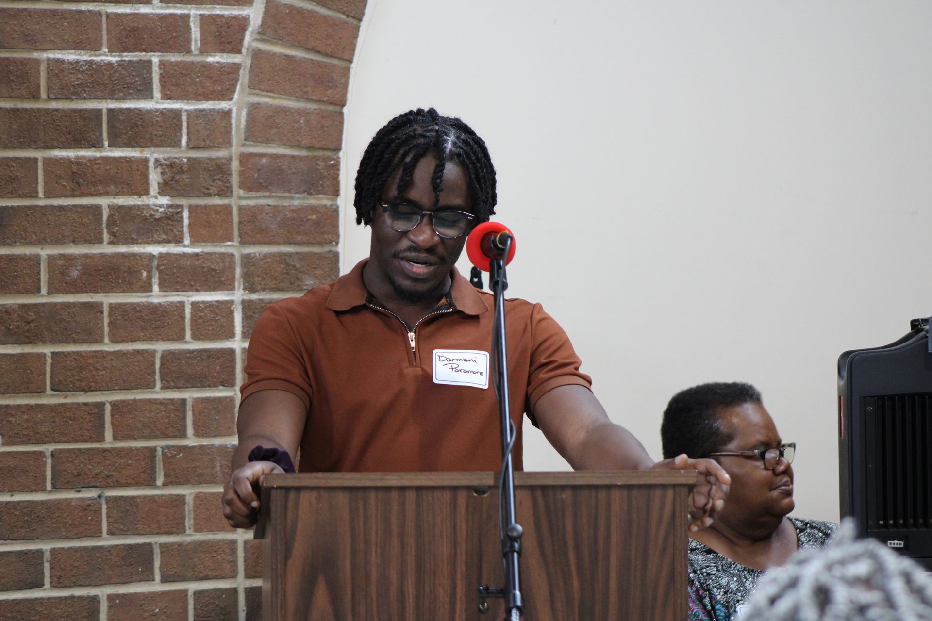 Man speaking at a podium with a microphone, in front of a brick wall and a seated person.