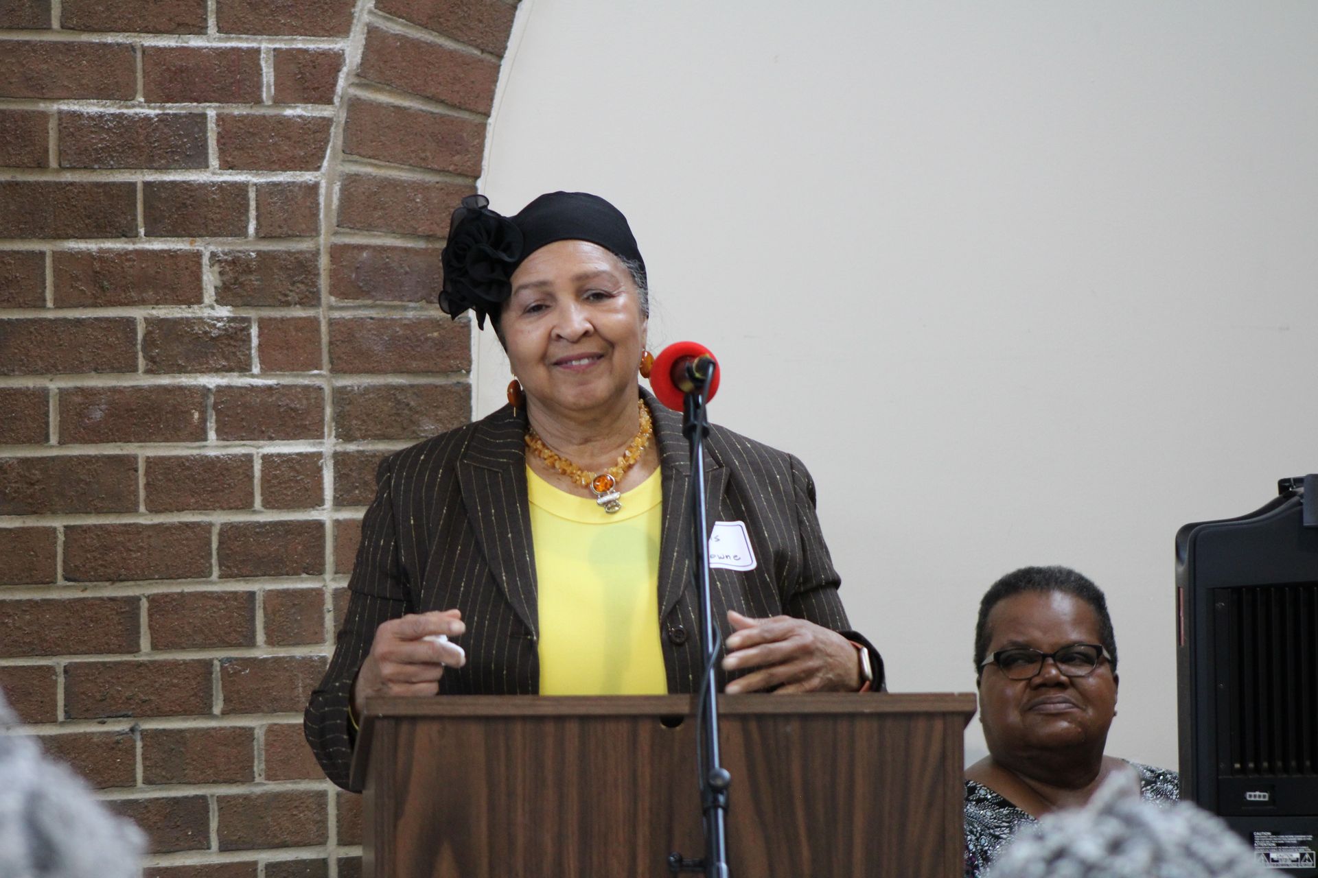 Woman speaking at a podium; brick wall background, another woman seated.
