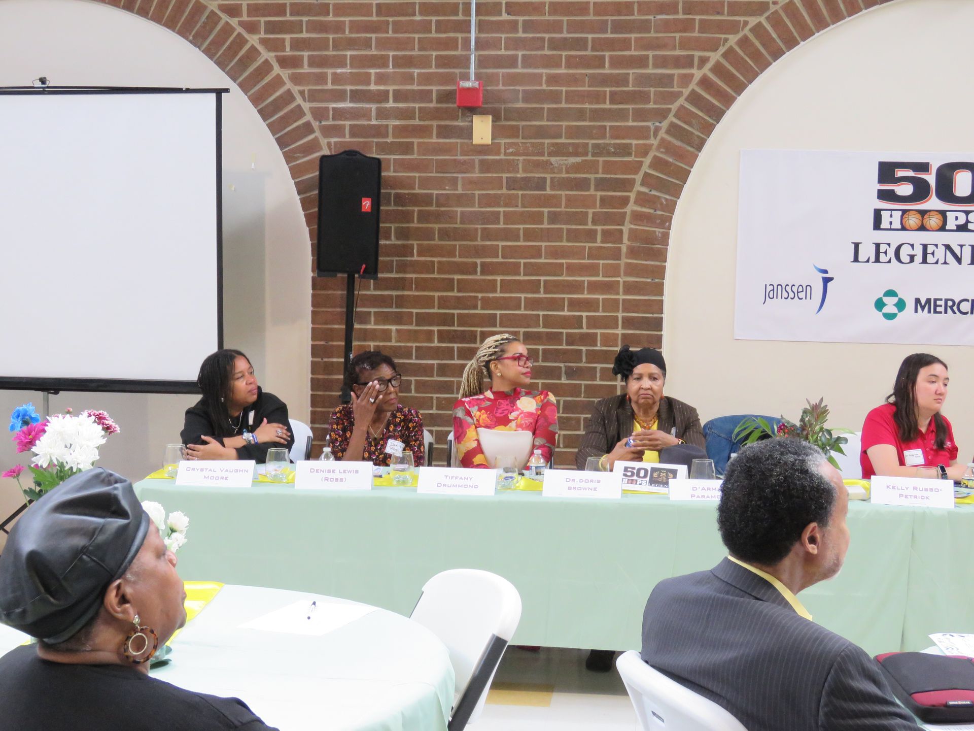 Panel of five women at a table, discussing topics. Audience members are seated in the foreground.