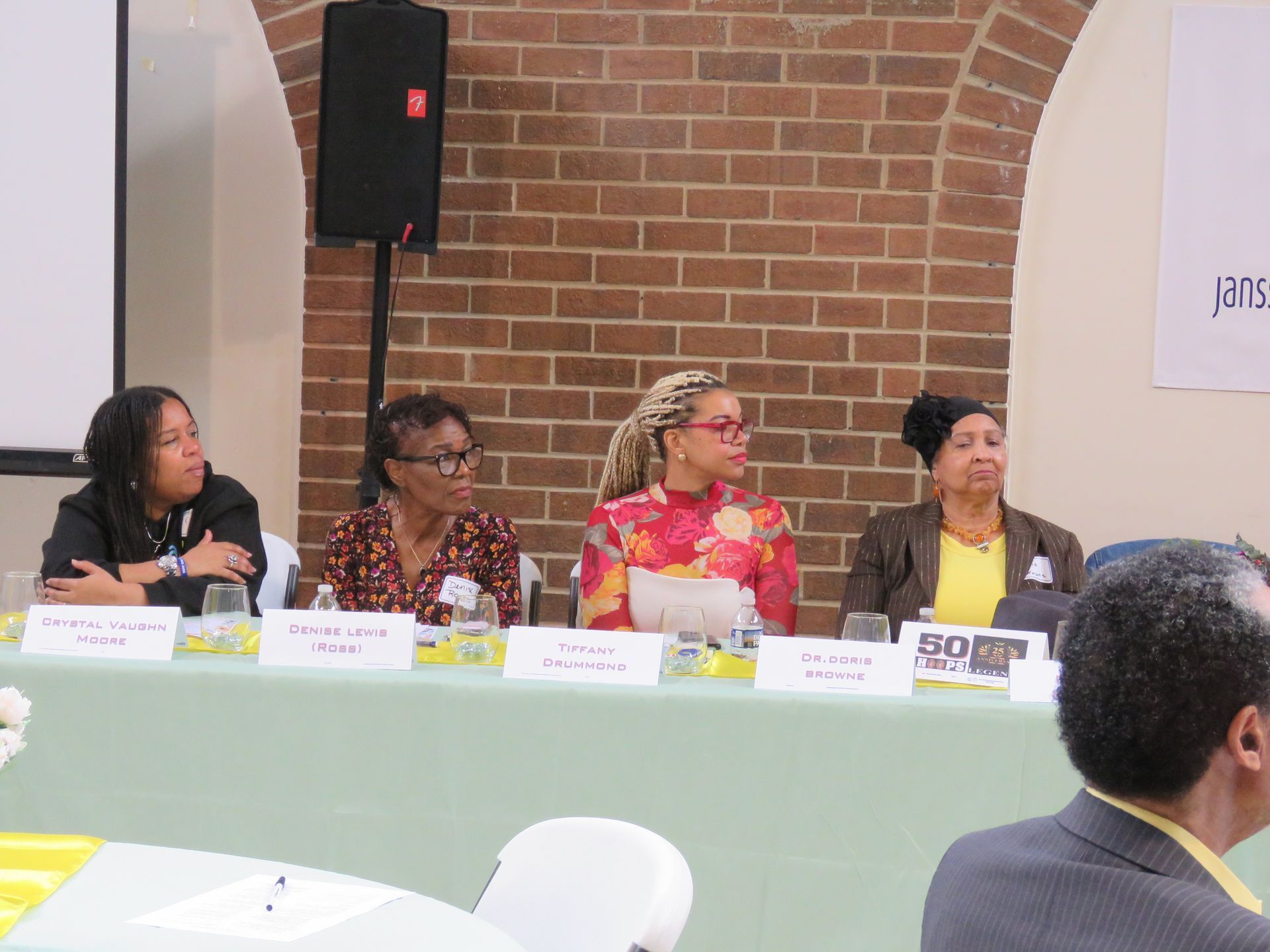 Panel of four speakers at a table; brick background.