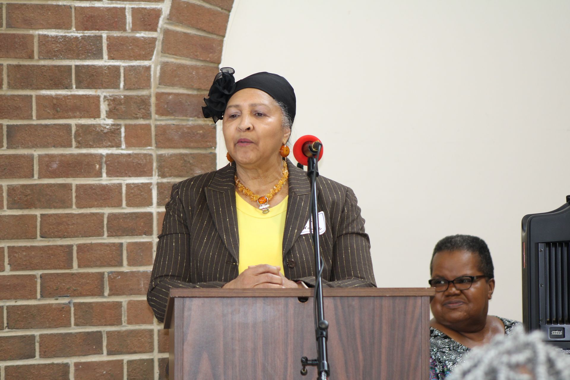 Woman speaking at a podium, brick wall and another person in the background.