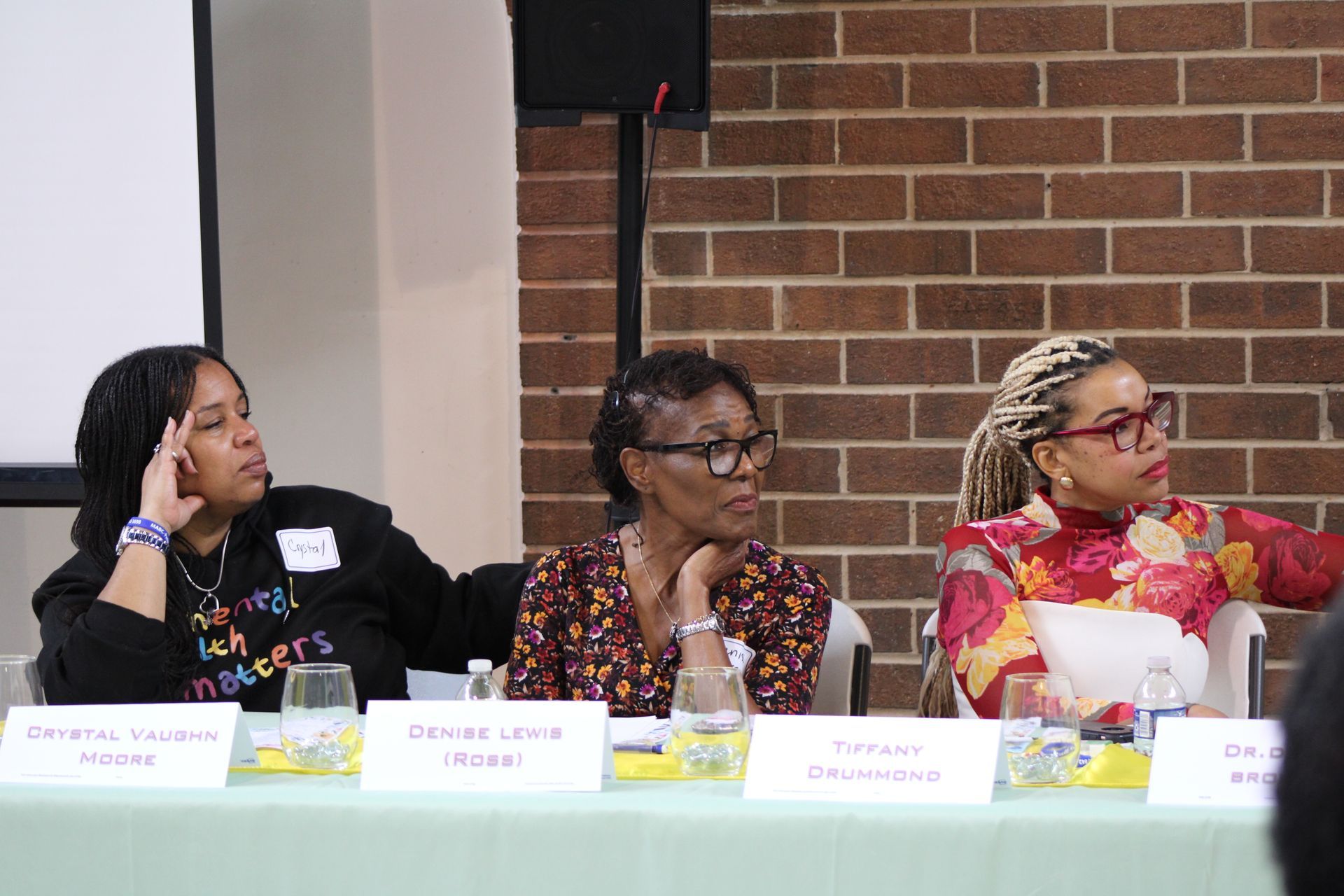 Three women at a panel discussion, seated behind a table, looking forward. Brick wall backdrop.