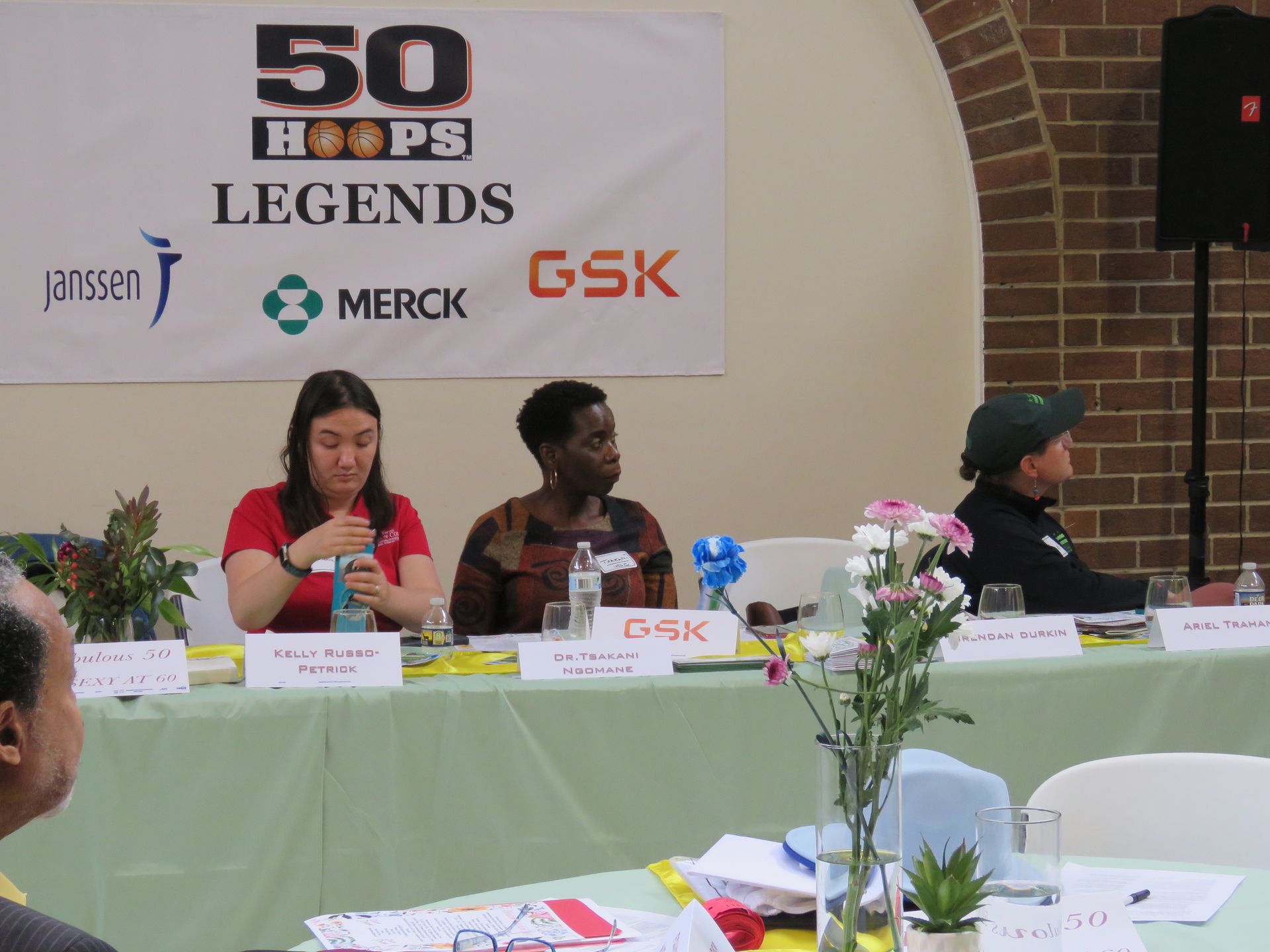 Panel with three speakers at a conference. They sit behind a table with water bottles and a banner with logos.