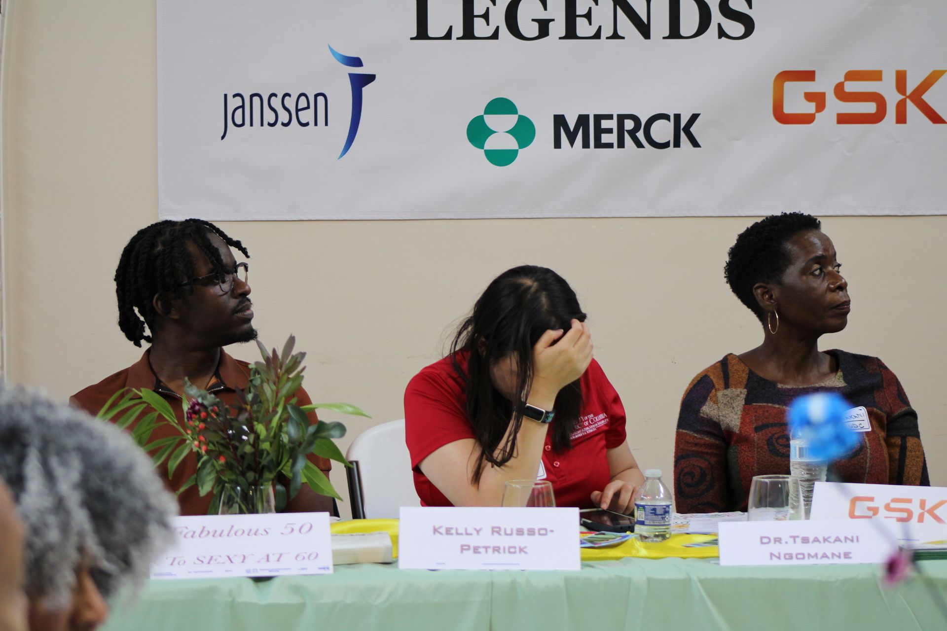 Three people at a table, facing forward. Middle person covers face. Sponsor logos overhead.