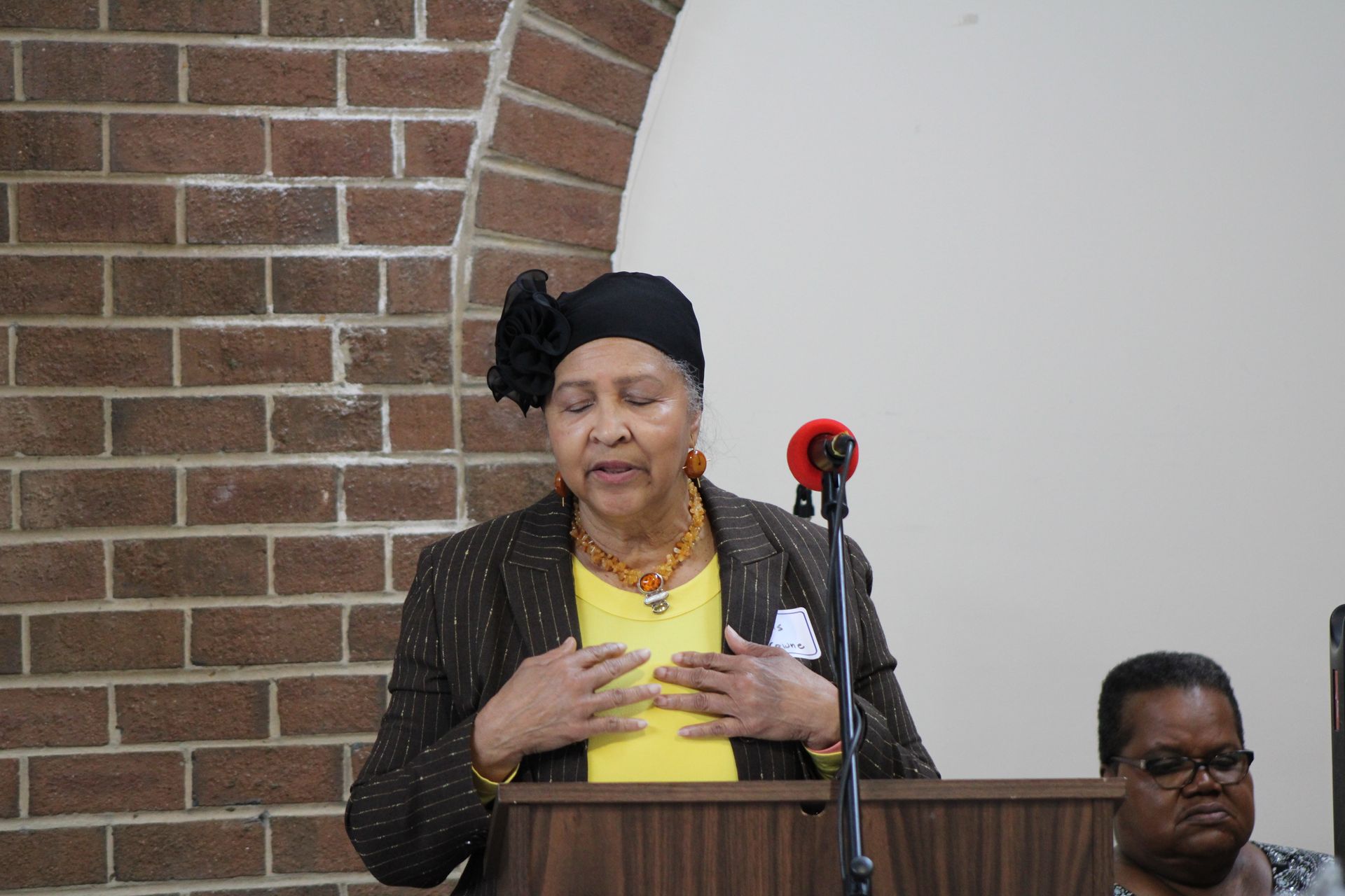 Woman speaking at a podium, wearing a black head covering and yellow shirt, hands on chest. Brick wall background.