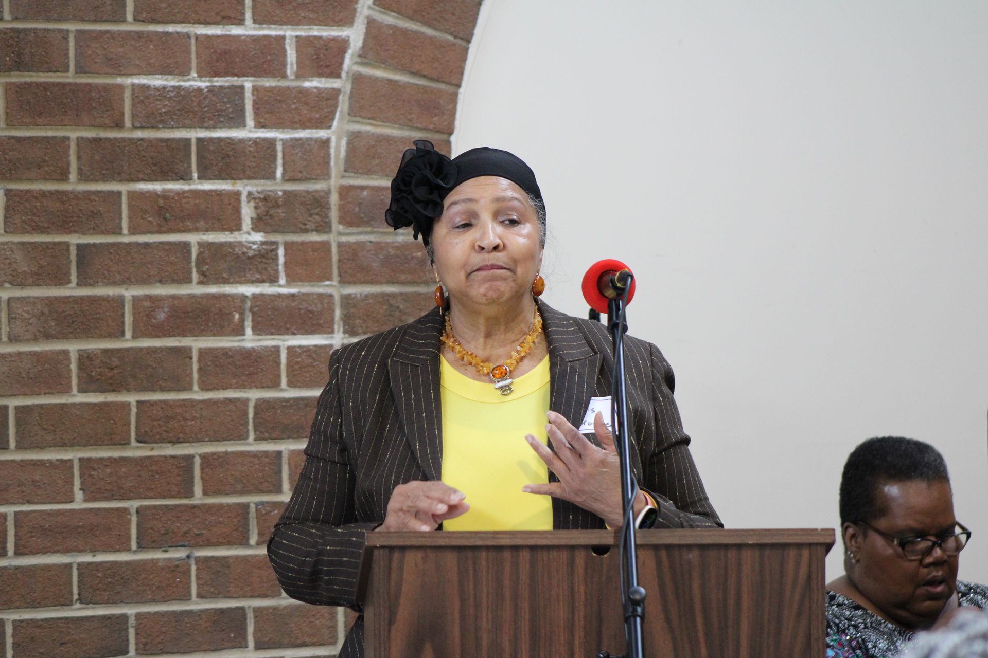 Woman speaking at podium, wearing black head covering and blazer, indoors near brick wall.
