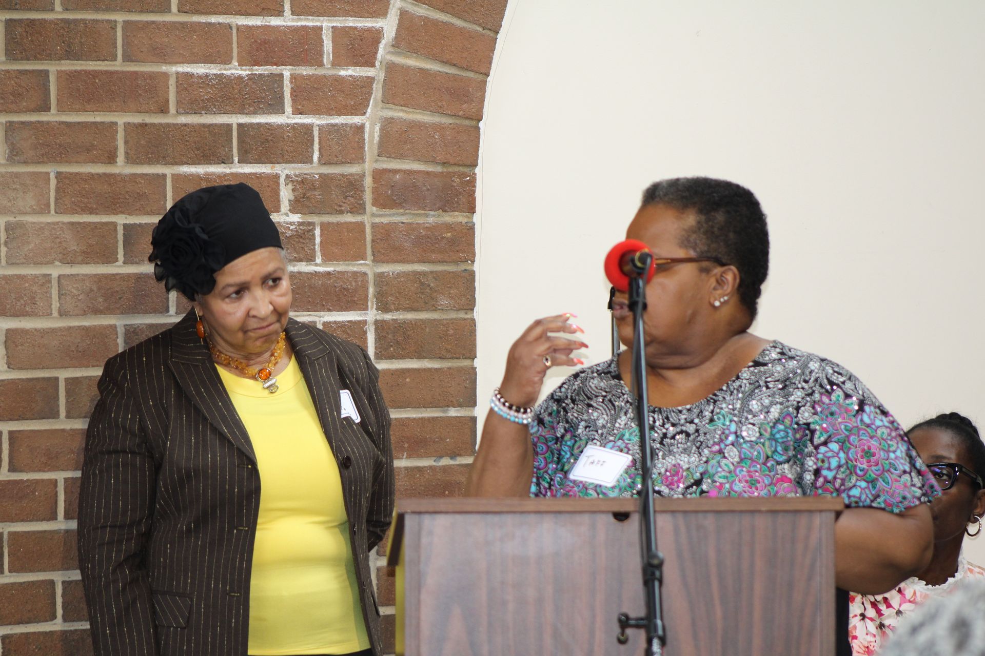 Woman speaking at podium, another woman beside her near brick wall.