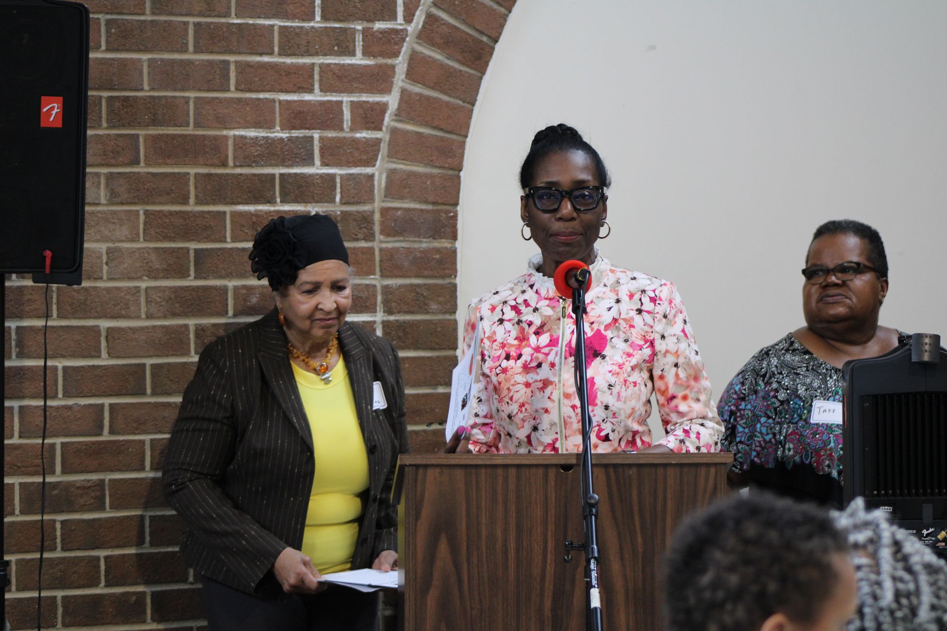 Three people at a podium, with a brick wall backdrop. One speaks into a microphone, while the others stand nearby.