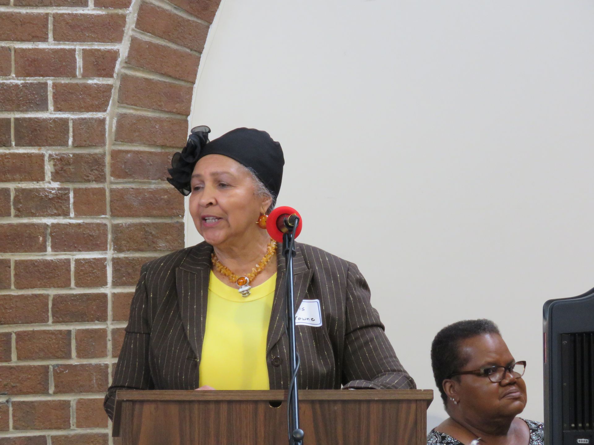 Woman speaking at a podium, wearing a dark hat and jacket, in a brick-walled room.