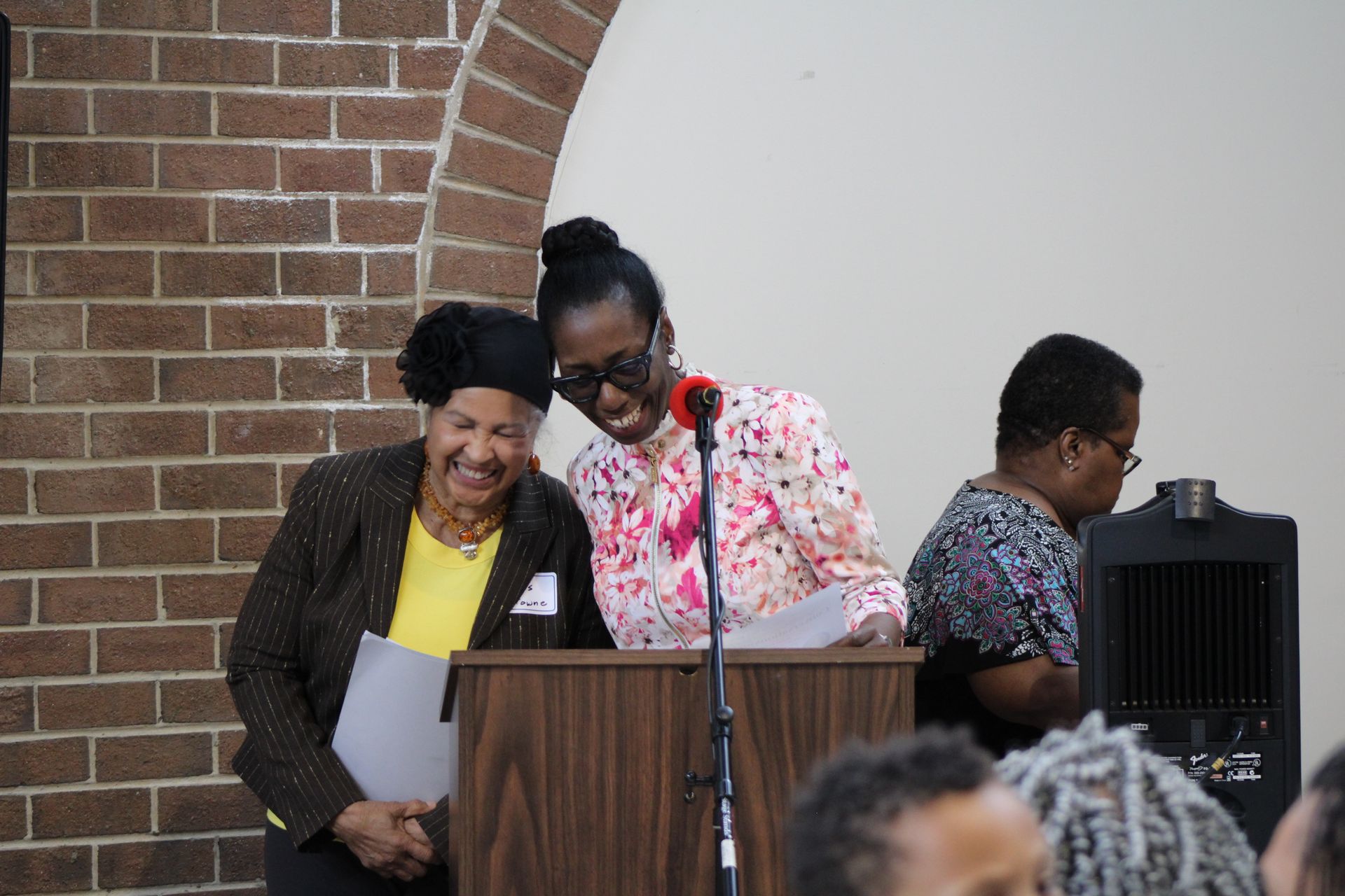 Two women smiling at a podium, papers in hand, brick wall background.