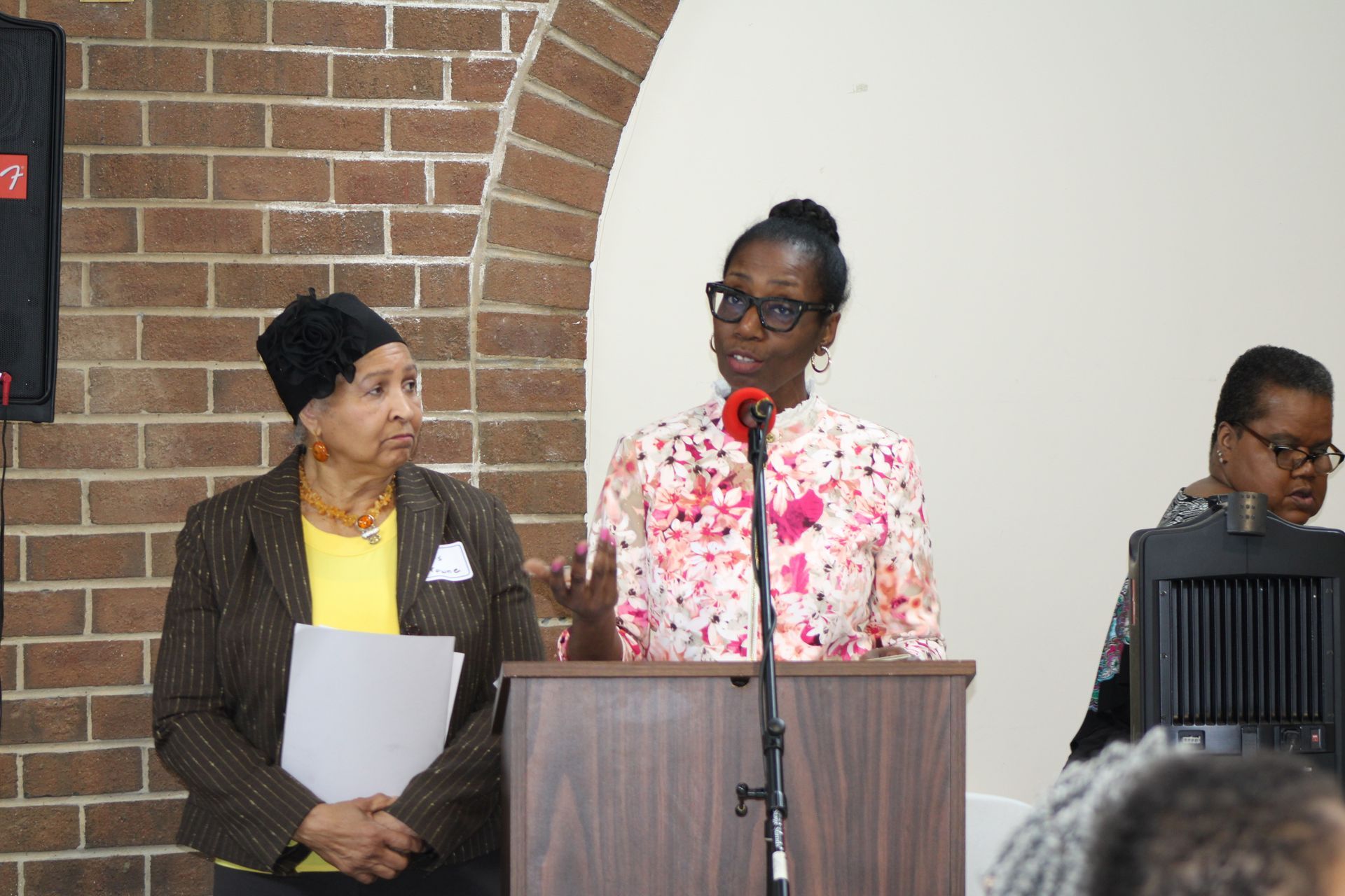 Woman speaking at podium, two others alongside, brick wall backdrop.