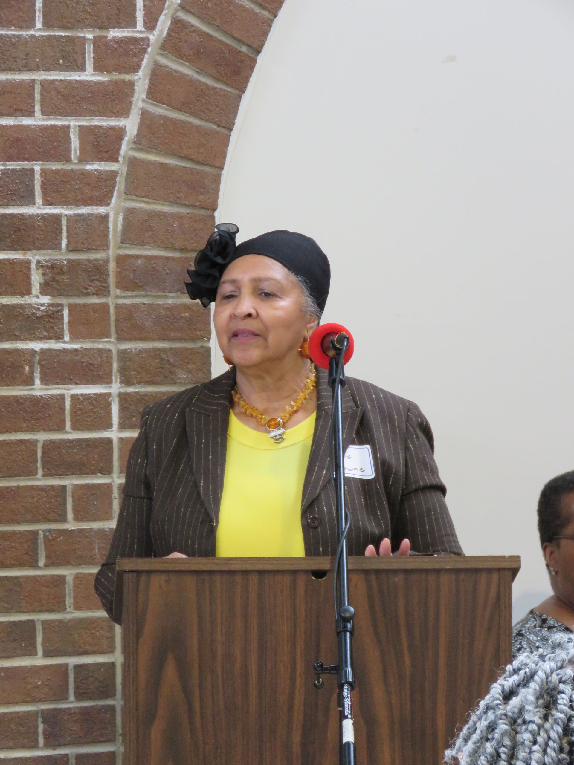 Woman speaking at a podium. She wears a yellow shirt, brown jacket, black head wrap, and jewelry. Brick arch backdrop.