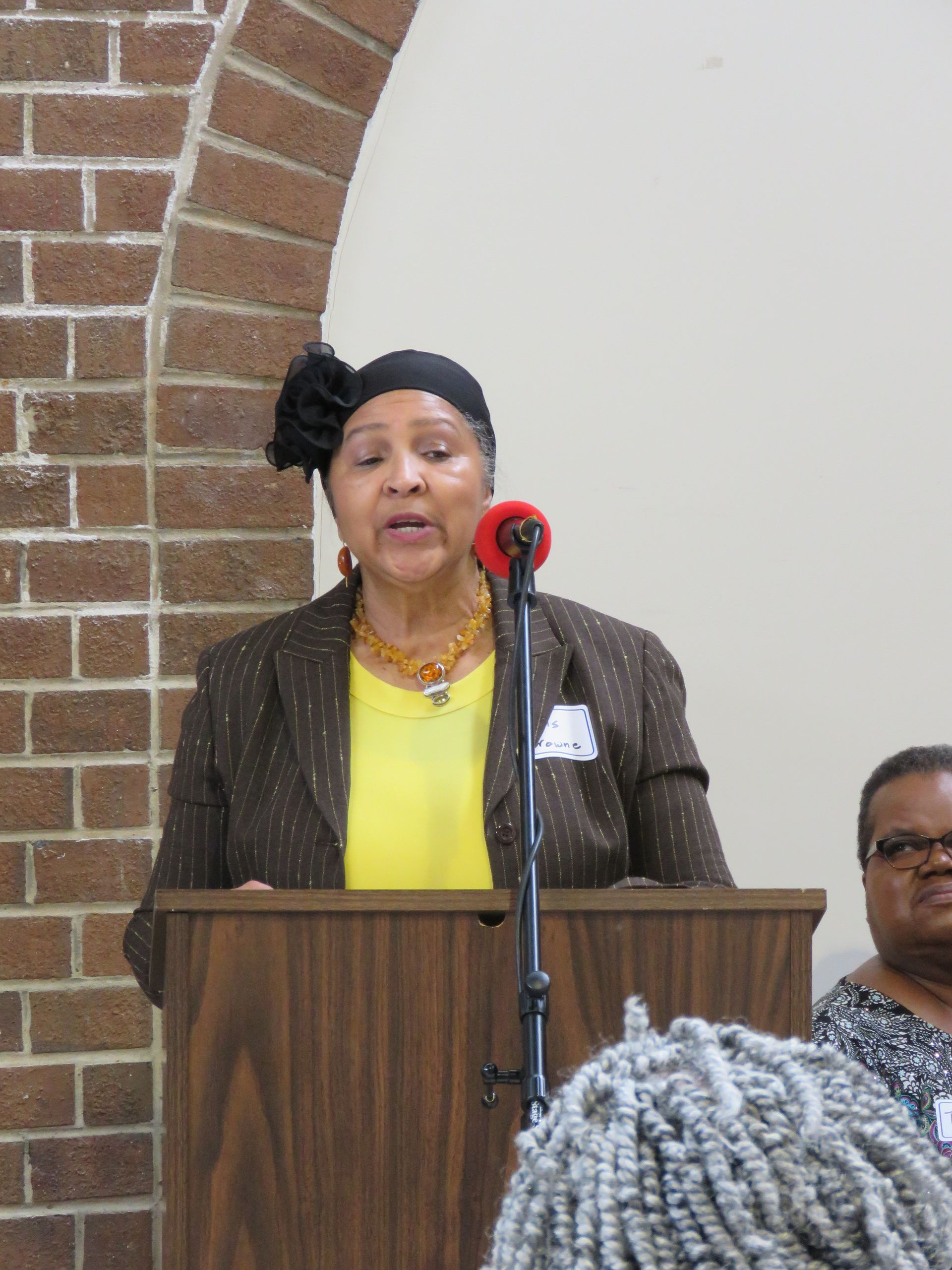 Woman speaking at a podium with a brick arch in the background. She wears a yellow shirt and brown jacket.