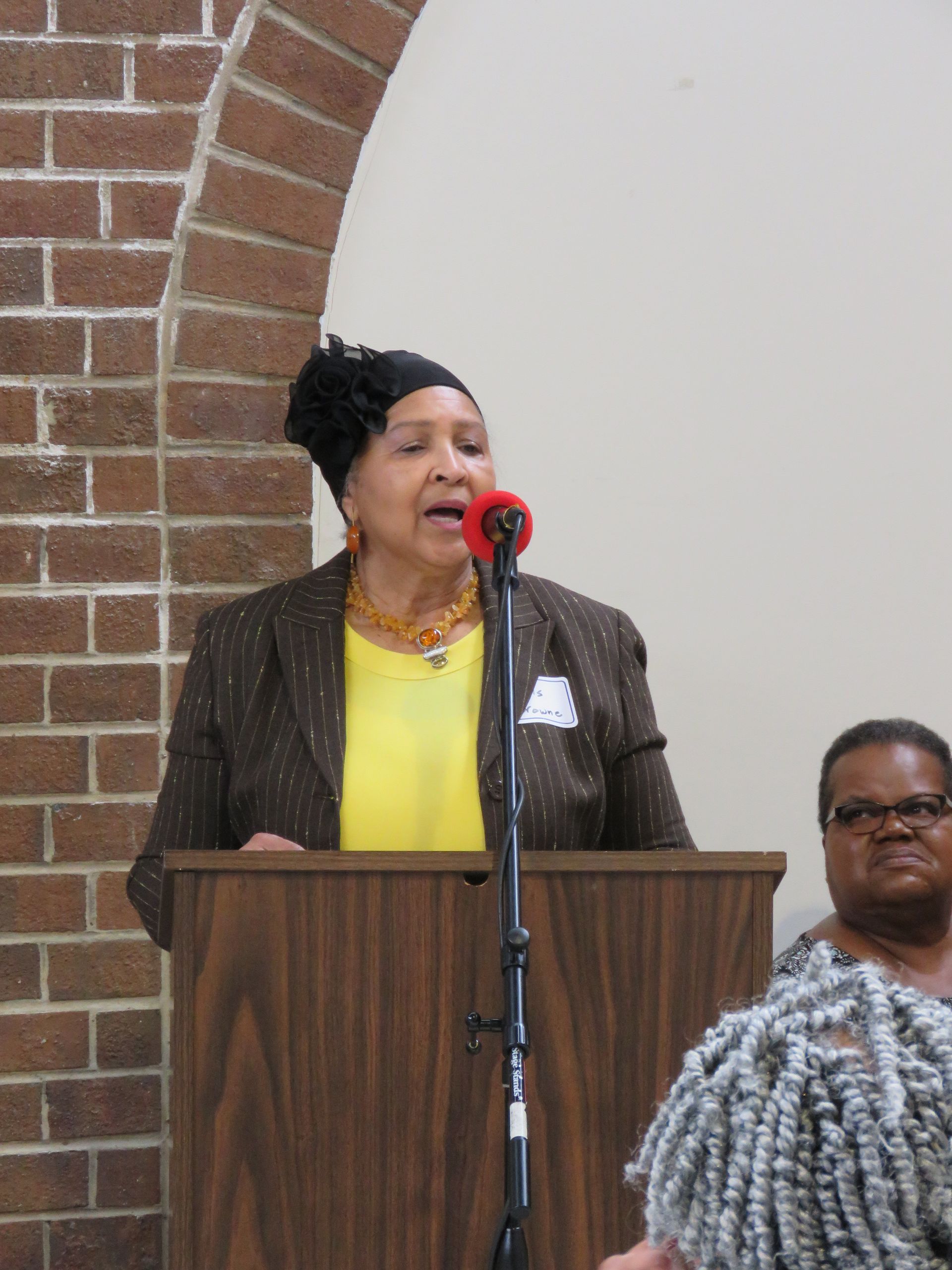 Woman speaking at a podium with microphone, wearing a blazer and yellow top; brick wall in background.