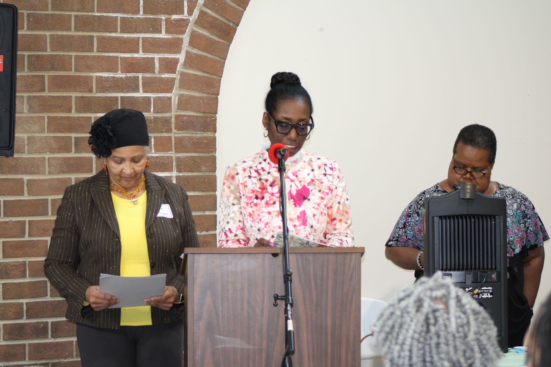 Three women speaking at a podium in front of a brick wall and a white wall.