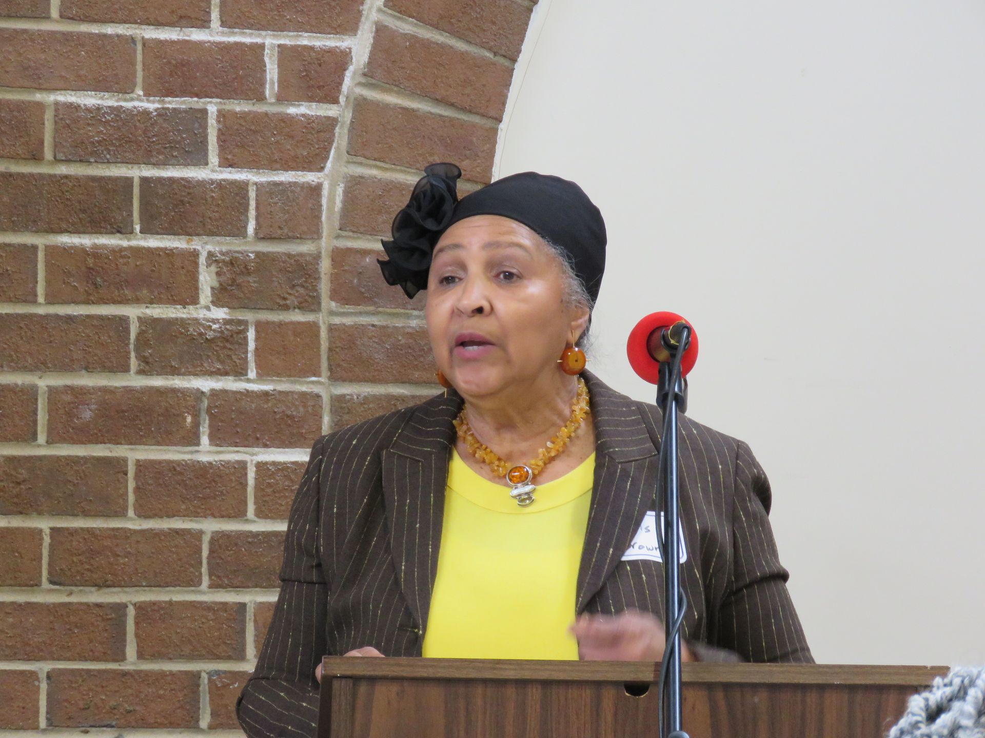Woman speaking at a podium in a brick-walled room; wearing black head covering, brown blazer, yellow shirt, and jewelry.