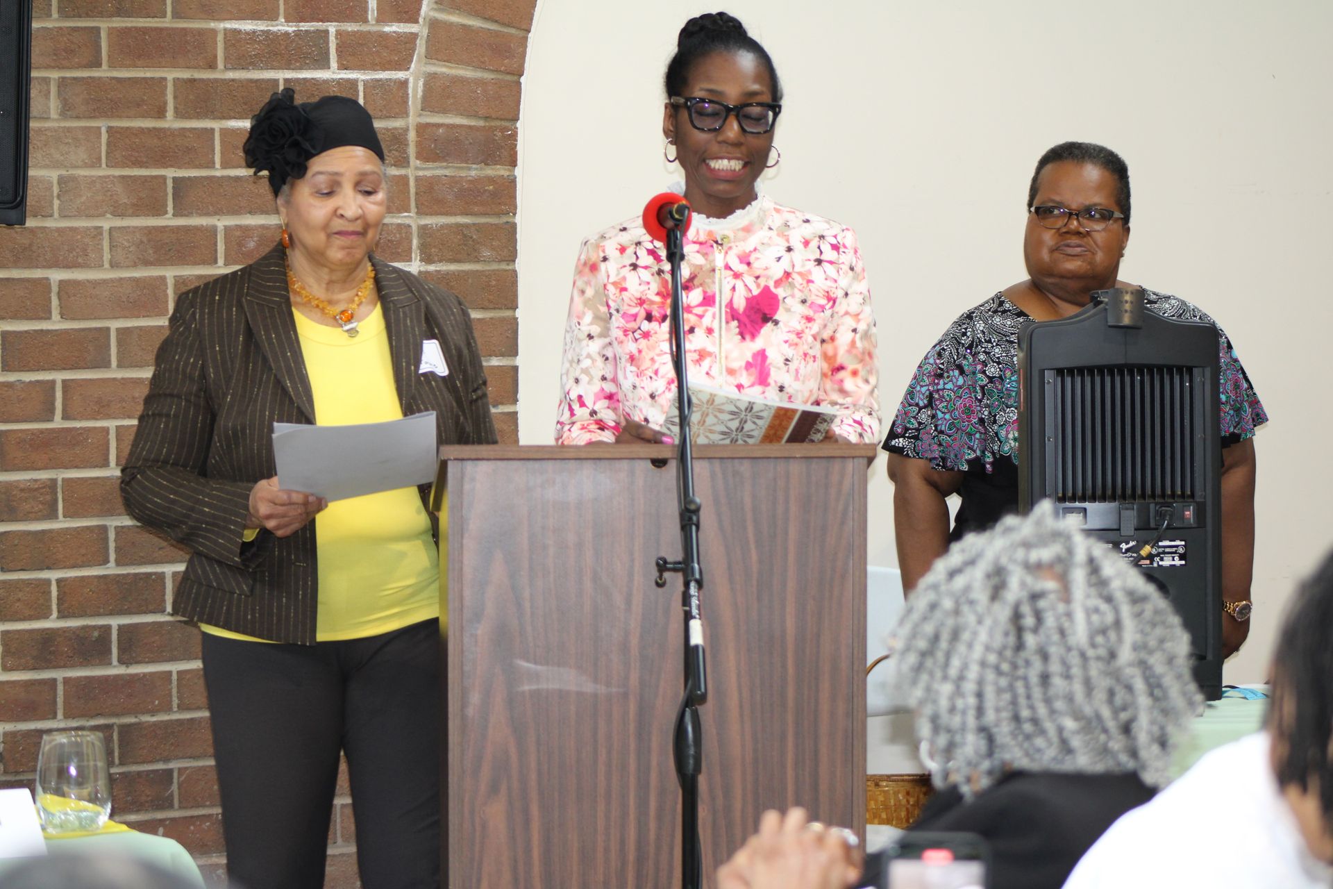 Three women stand at a podium, speaking. One holds paper, two look forward. Interior setting.