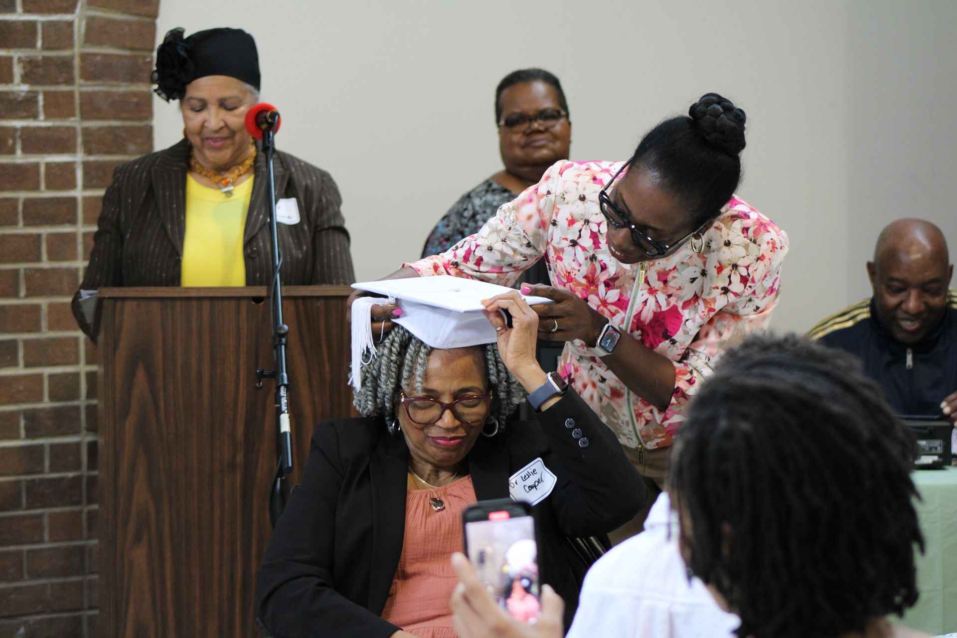 Woman being presented with a graduation cap at a podium. Others look on, one taking a photo.