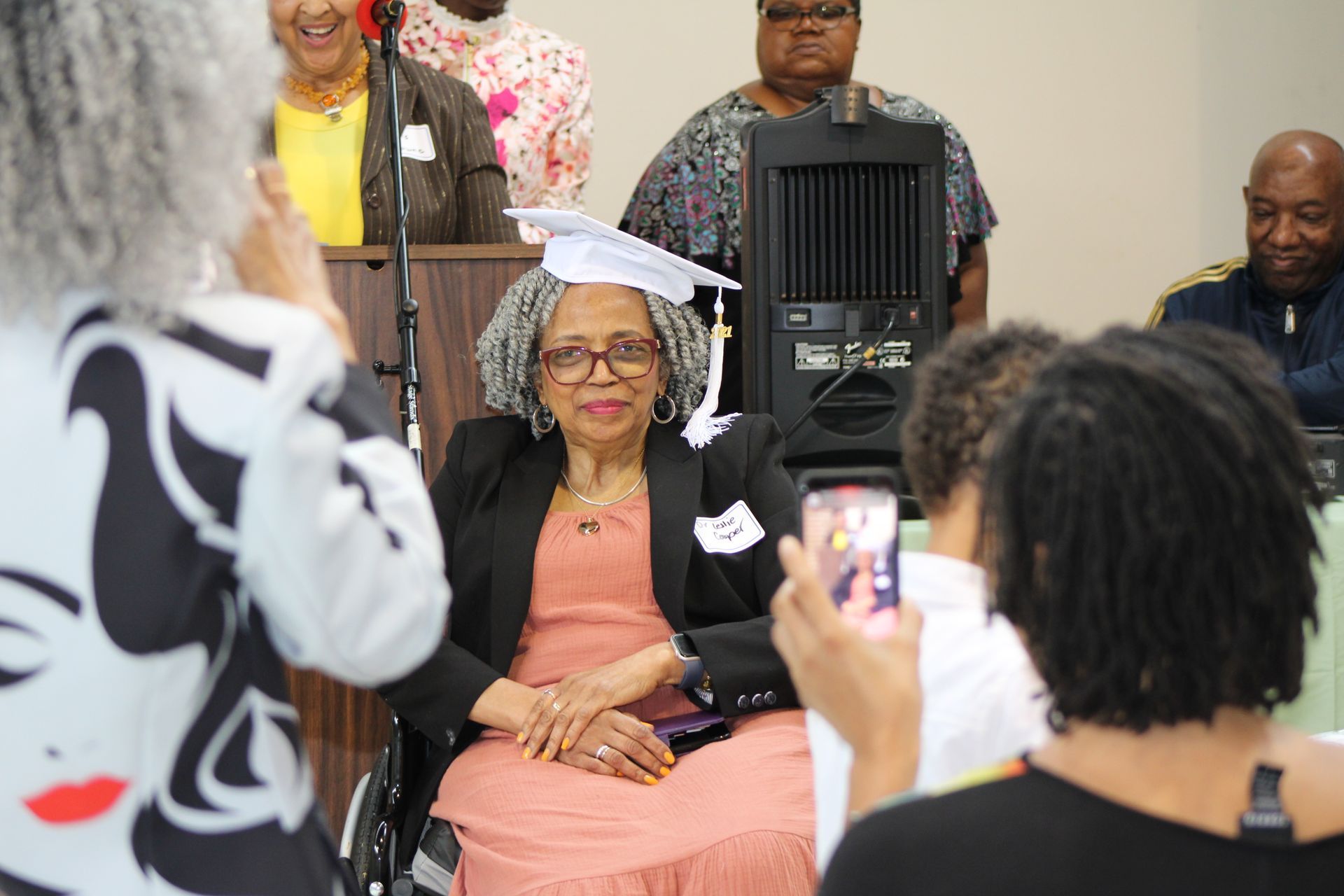 Woman in cap and gown, smiling, being photographed at a ceremony. Others in background.