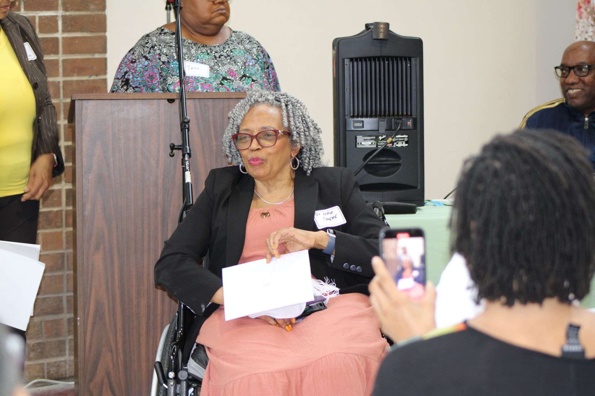 Woman in wheelchair speaks at podium, holding papers. Other people seated around table.