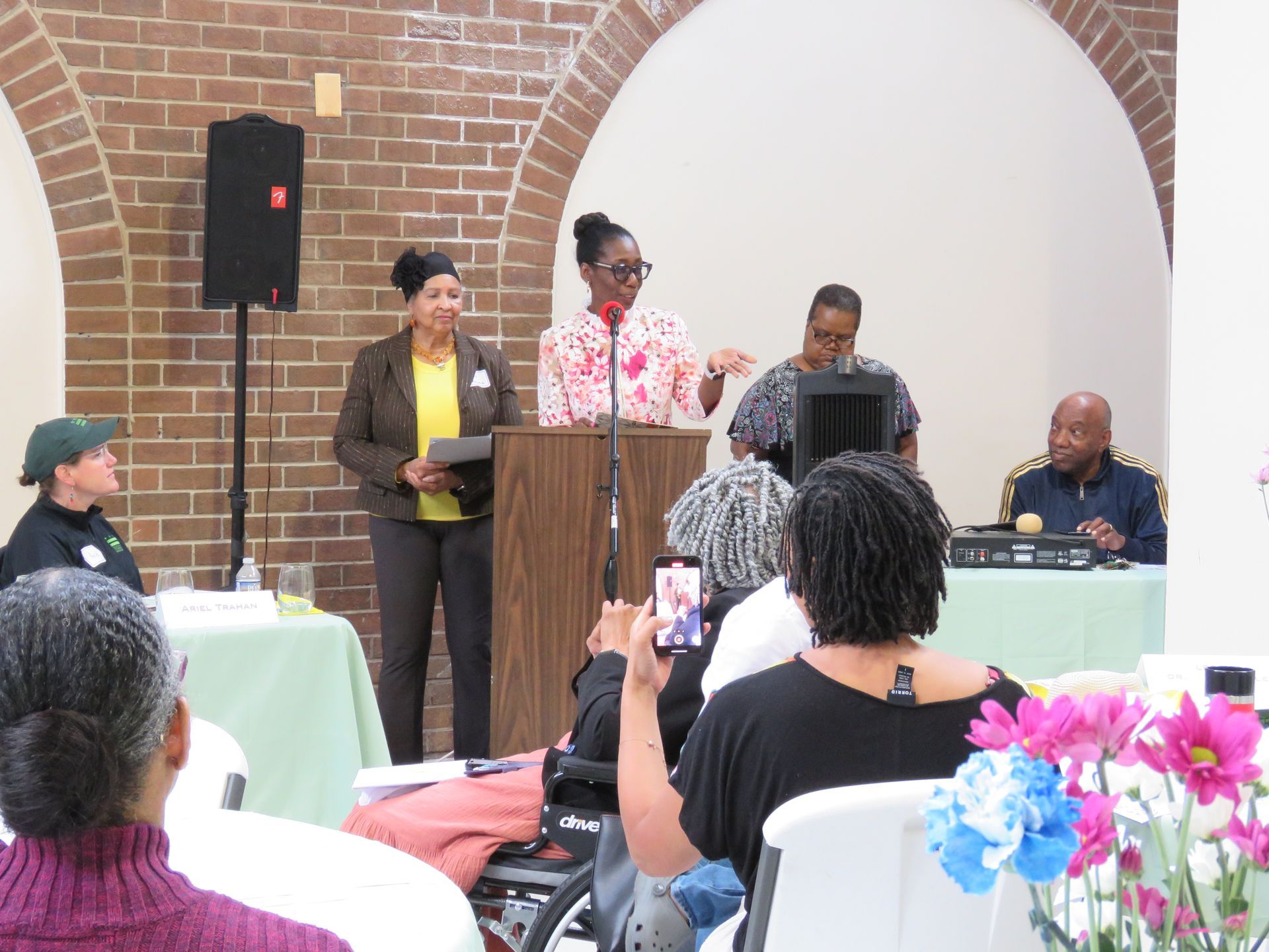People at a podium, speaking at a public meeting. Audience seated at tables with flowers. Brick wall backdrop.