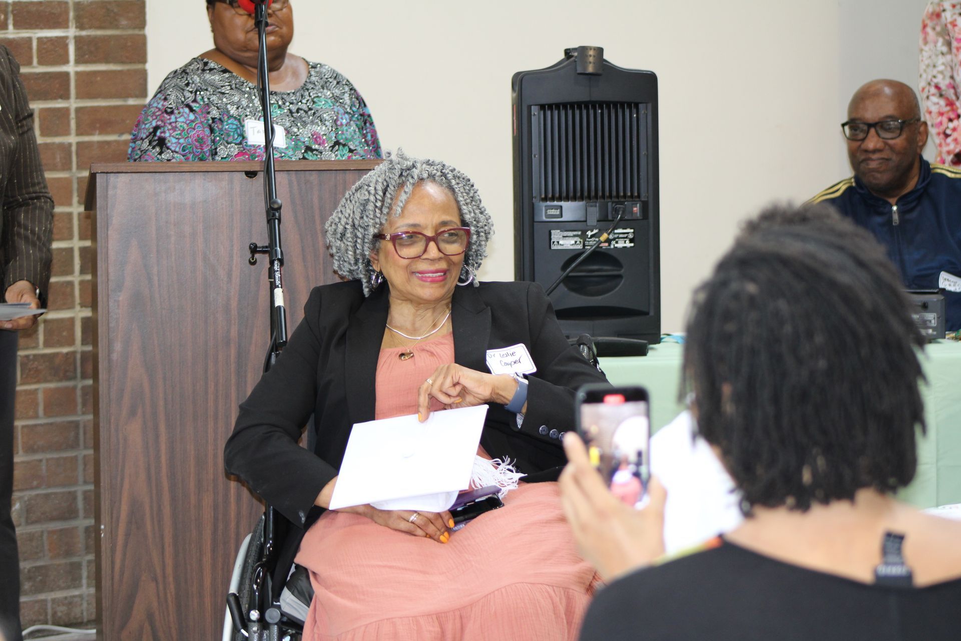 Woman in wheelchair, holding papers, smiles. Someone takes a photo. Others seated at table and podium.