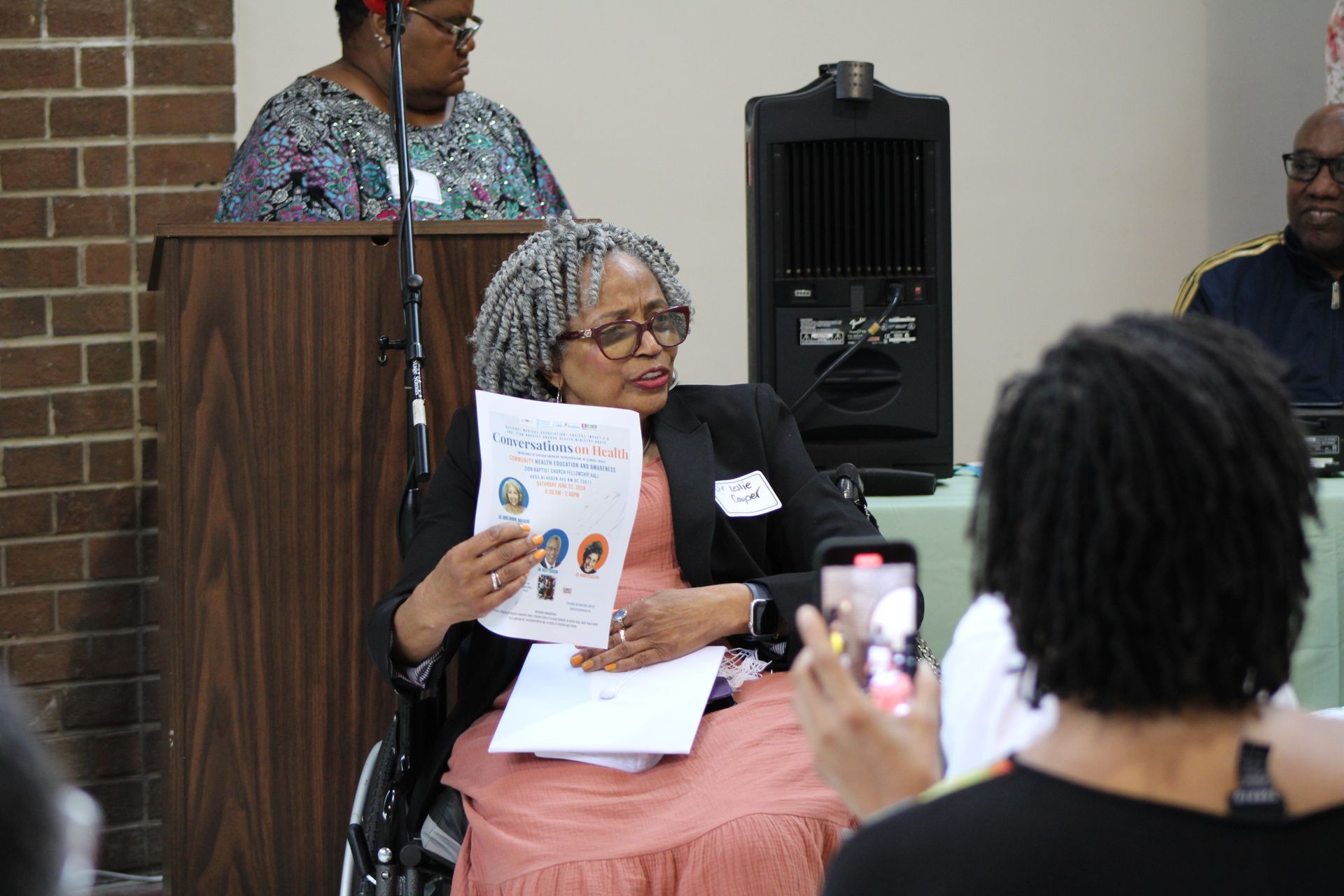 Woman in wheelchair presenting paper to a person with a phone; a lectern, speaker, and people are behind her.