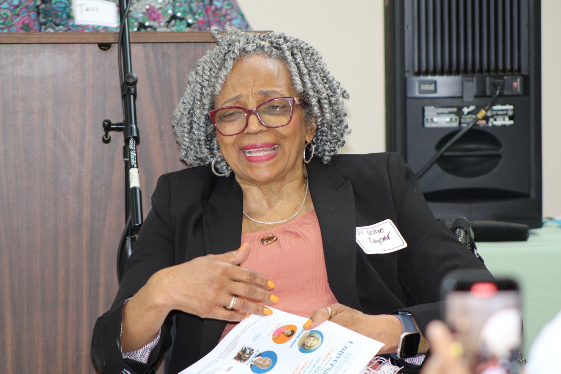 Woman in black blazer speaks, holding paper; seated by speaker, microphone.