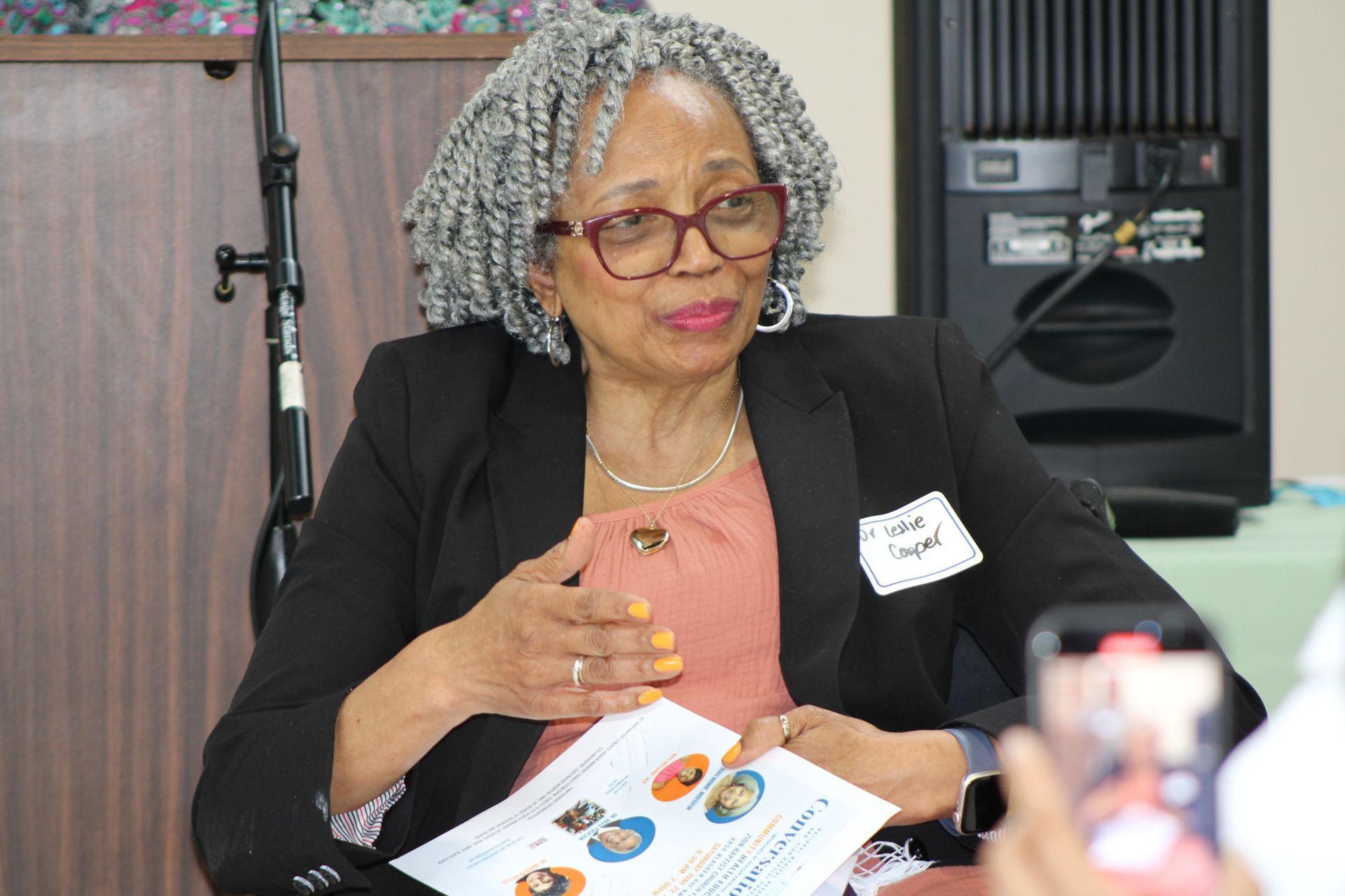 Woman with gray hair and glasses speaking, holding a paper; she wears a black jacket and is indoors.