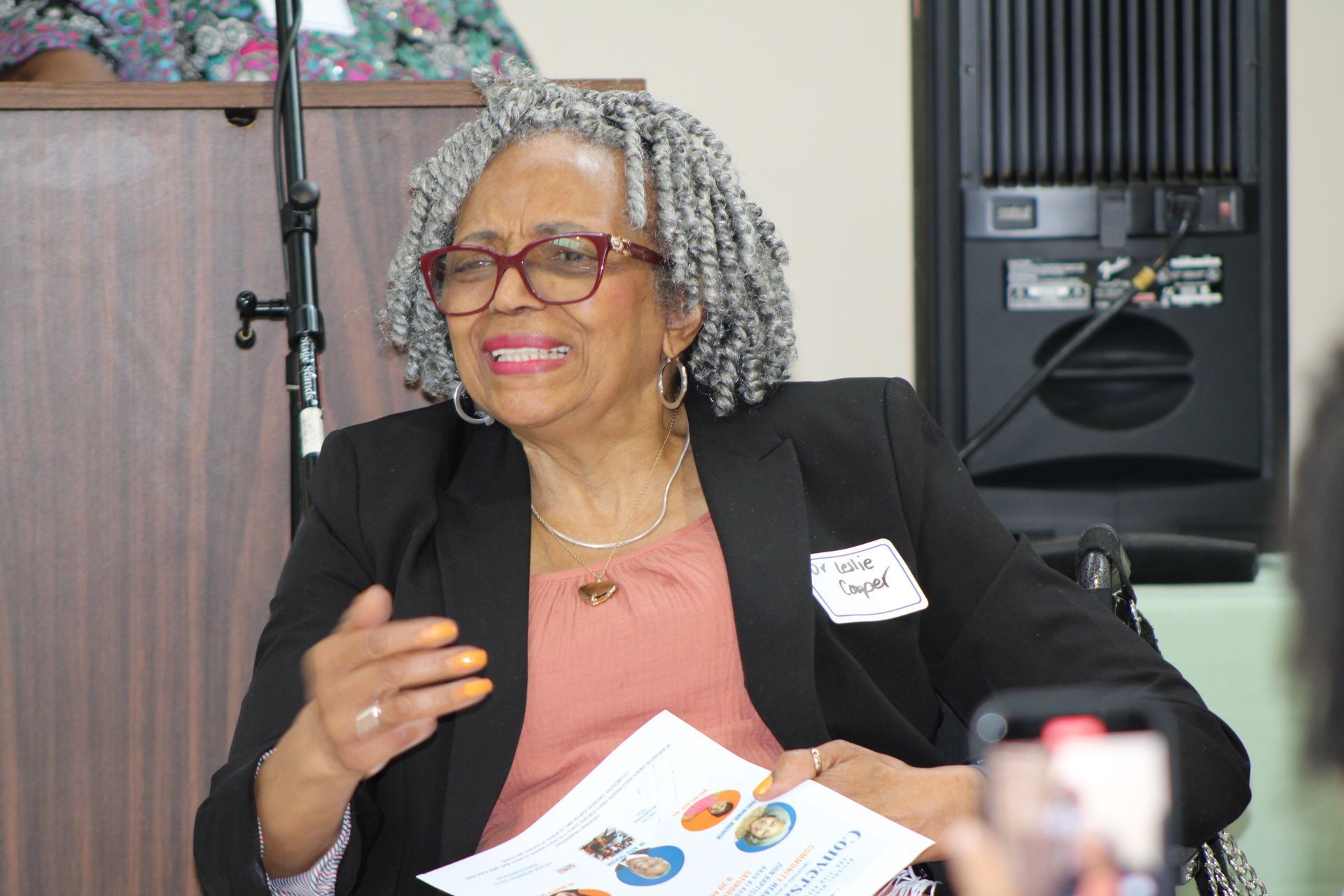 Woman in glasses, black blazer, smiles while gesturing, seated near podium and speaker.