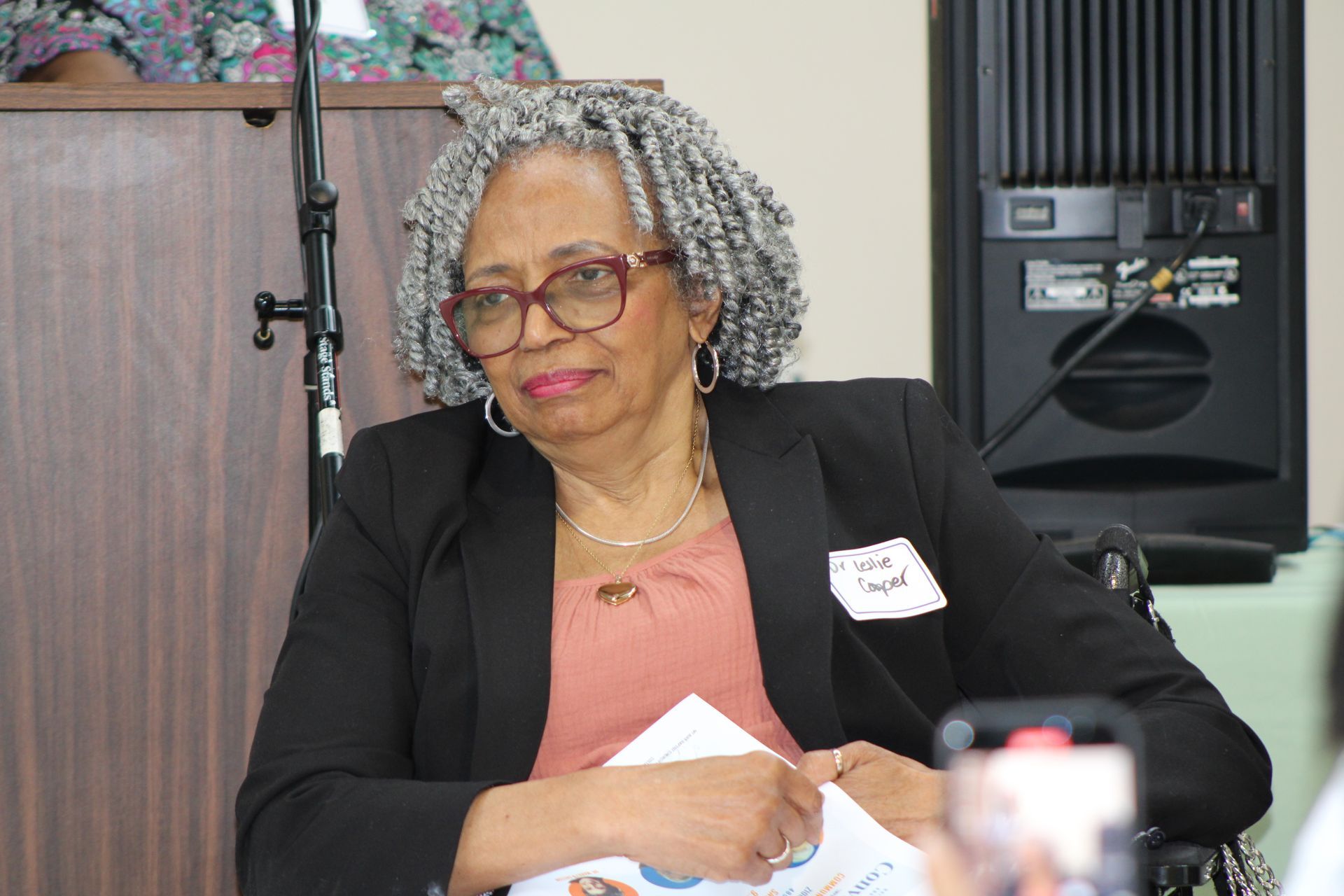 Woman with gray curly hair, wearing glasses and a black blazer, sitting indoors.