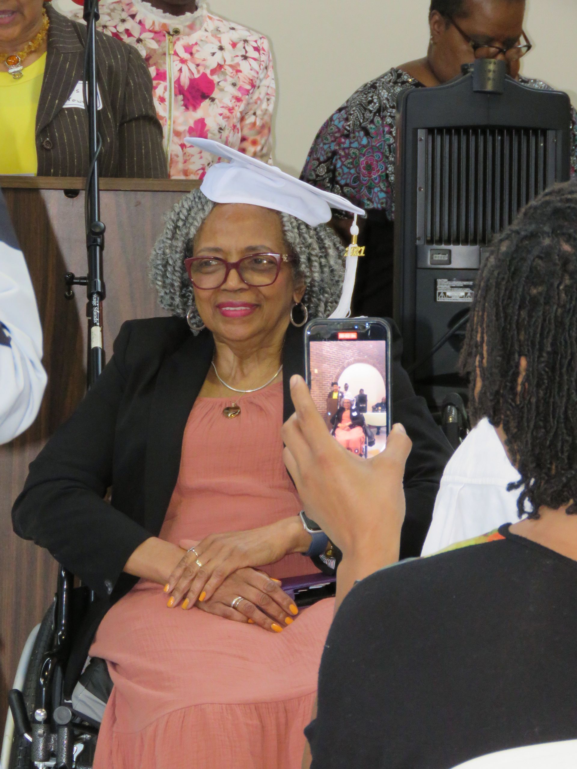 Woman in a wheelchair wearing a graduation cap, being photographed. She's smiling in a room.