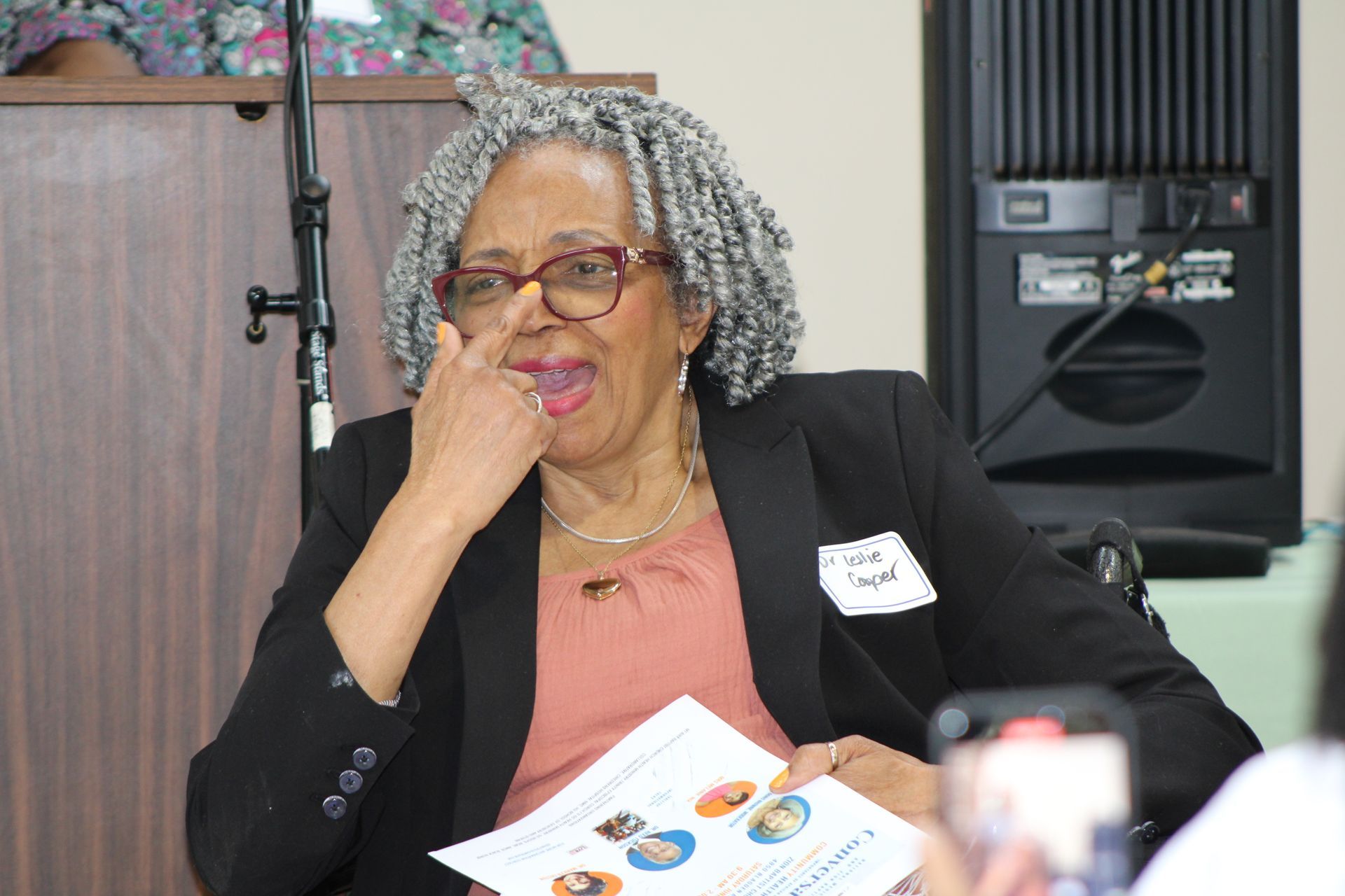Woman in a black blazer adjusts her glasses while seated, holding a paper. Speaker, audio equipment behind her.