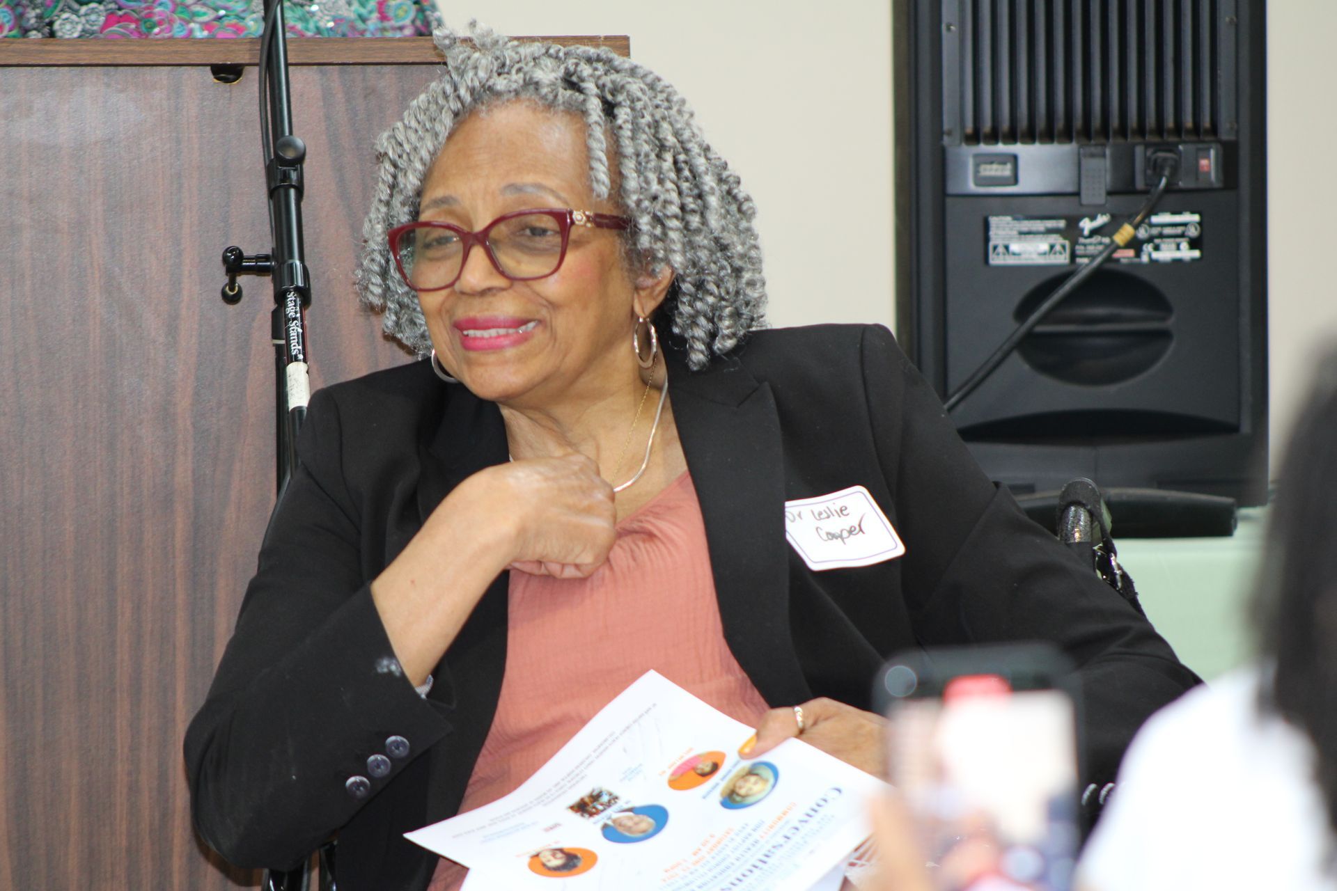 Woman with glasses and grey hair, wearing a blazer, holding papers and smiling at an event.