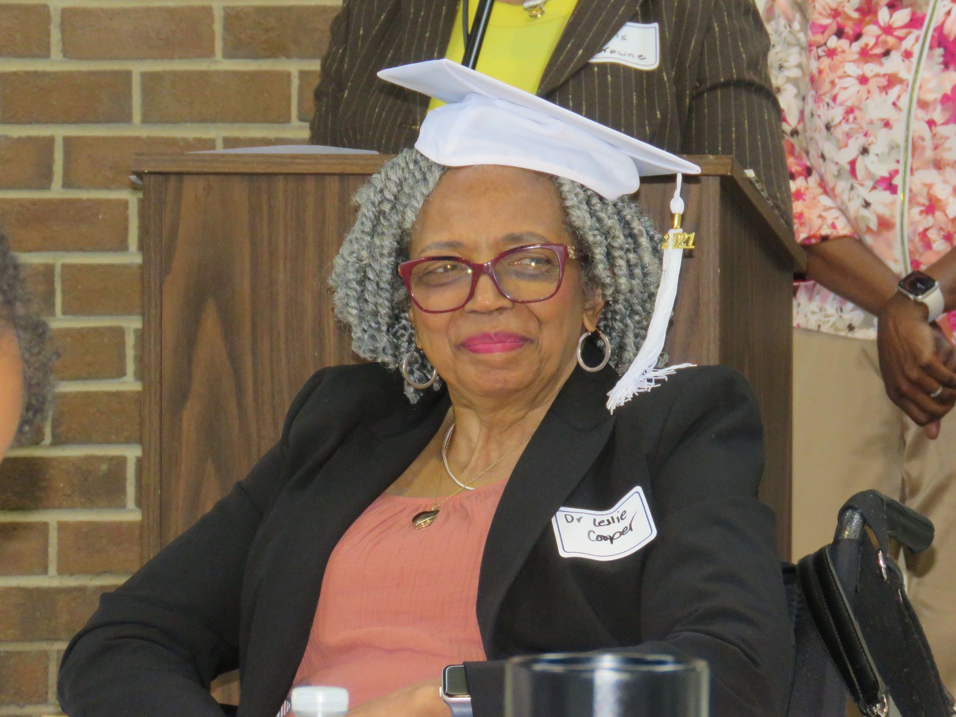 Woman with glasses and graduation cap, smiles at a podium. She wears a black blazer and peach top.