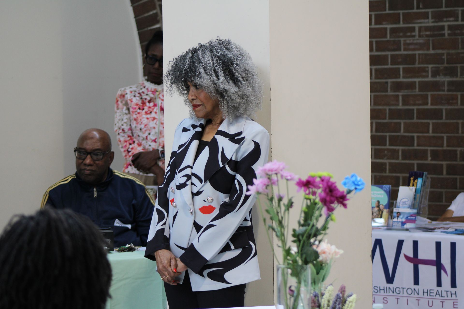 Woman in graphic jacket speaks at event; others listen. Flowers in foreground.