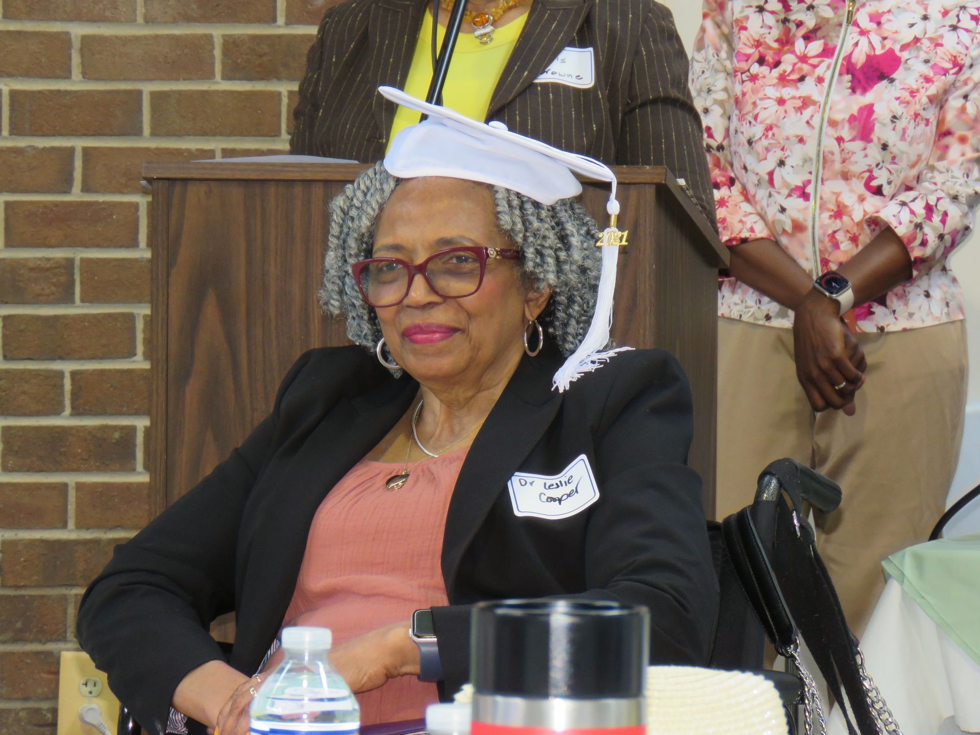 Woman in cap and gown, smiling, seated in a wheelchair, at podium, surrounded by people.