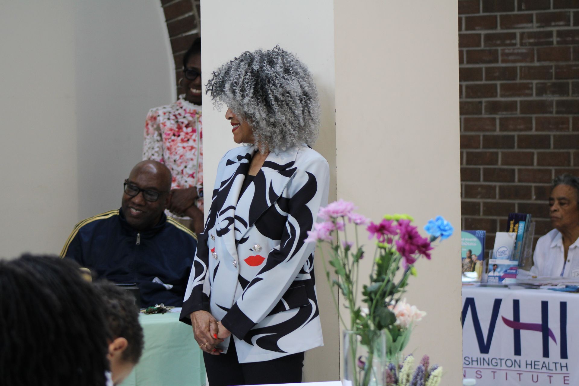 Woman in black and white jacket smiles, next to flowers and people at the Washington Health Institute event.