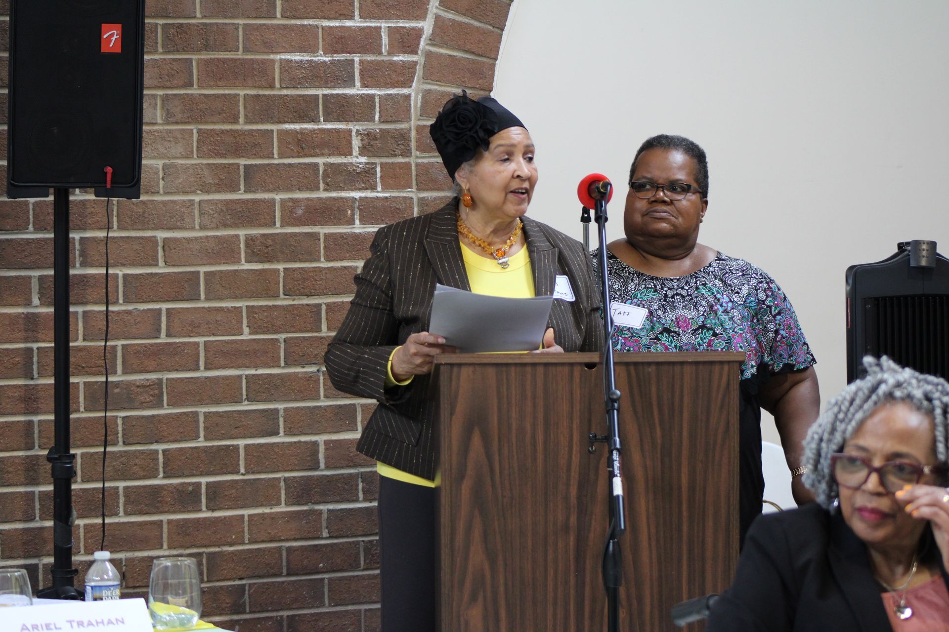Woman at podium speaking, another woman stands behind her. Brick wall in background.