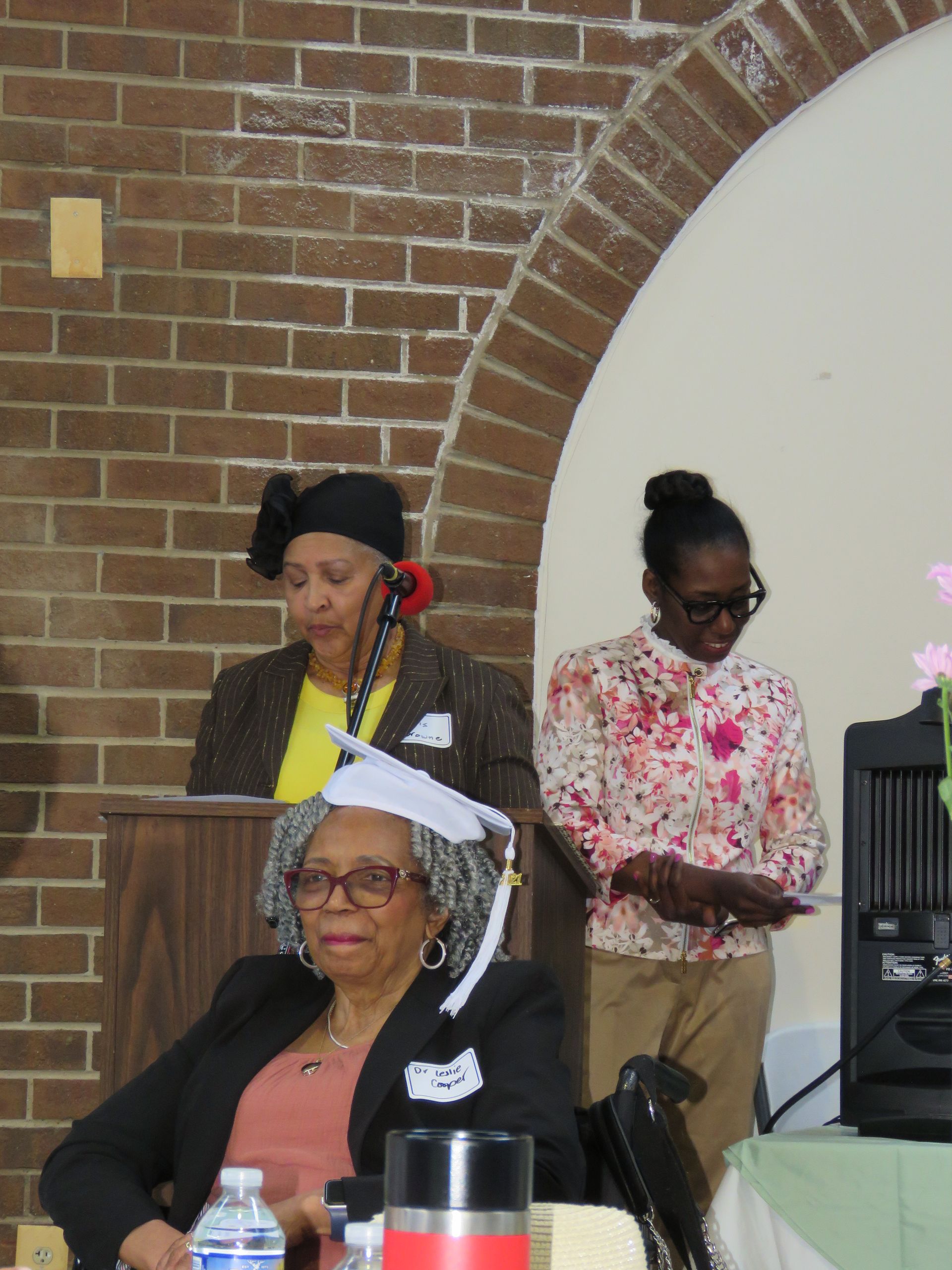 Three women at podium and table; brick archway. One seated with cap, two standing with microphones.