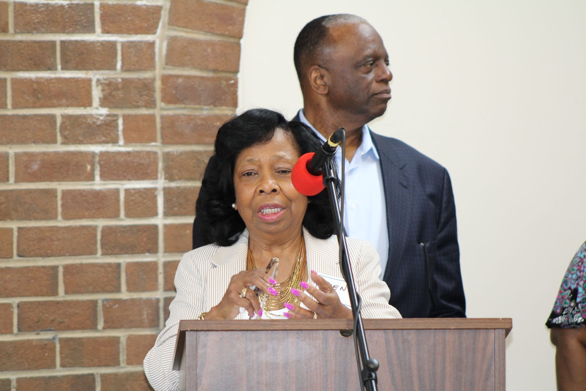 Woman speaking at a podium, man standing behind her, brick wall background.