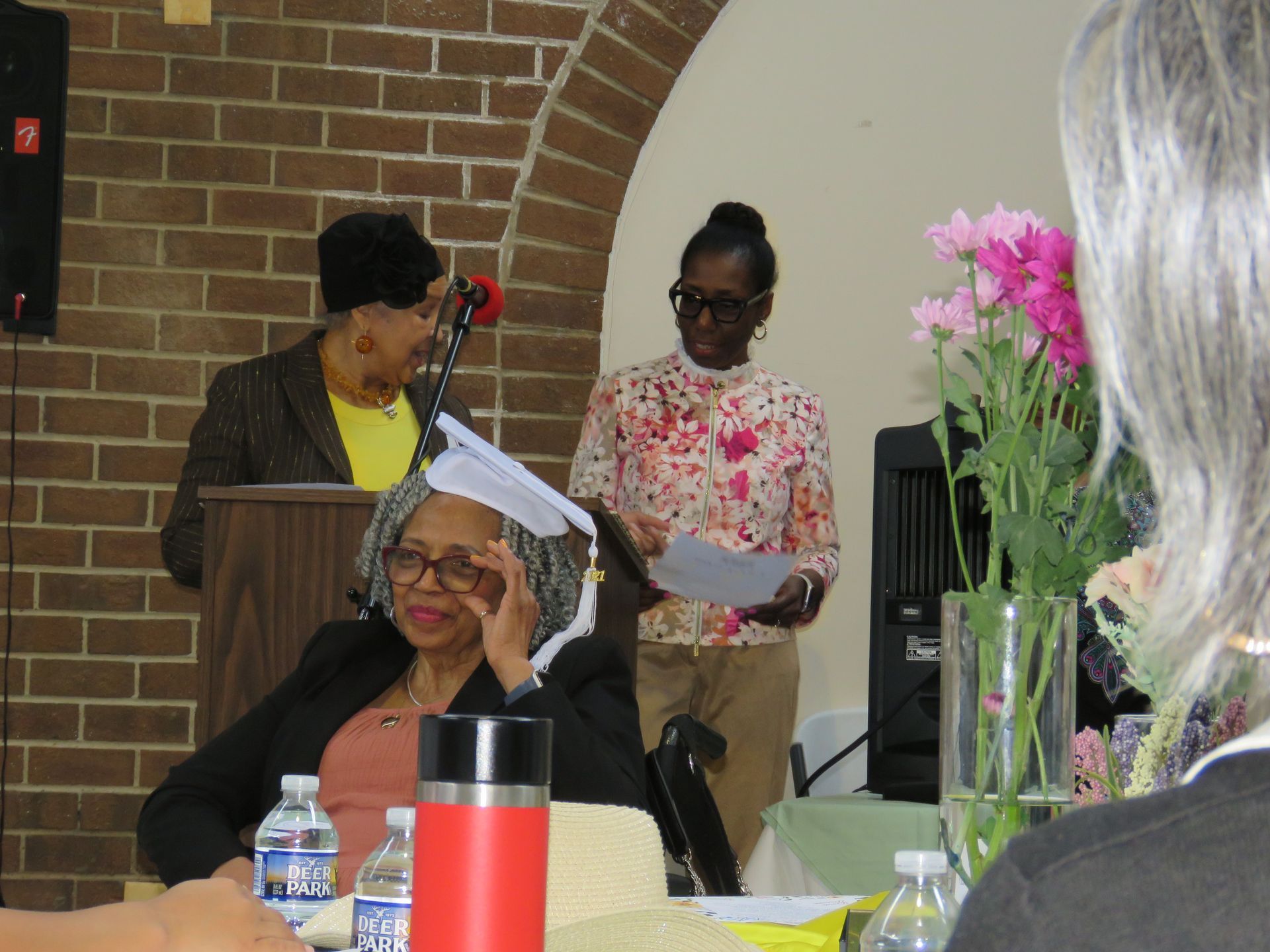 Woman in graduation cap sits at table, others stand and speak near brick arch and flowers.