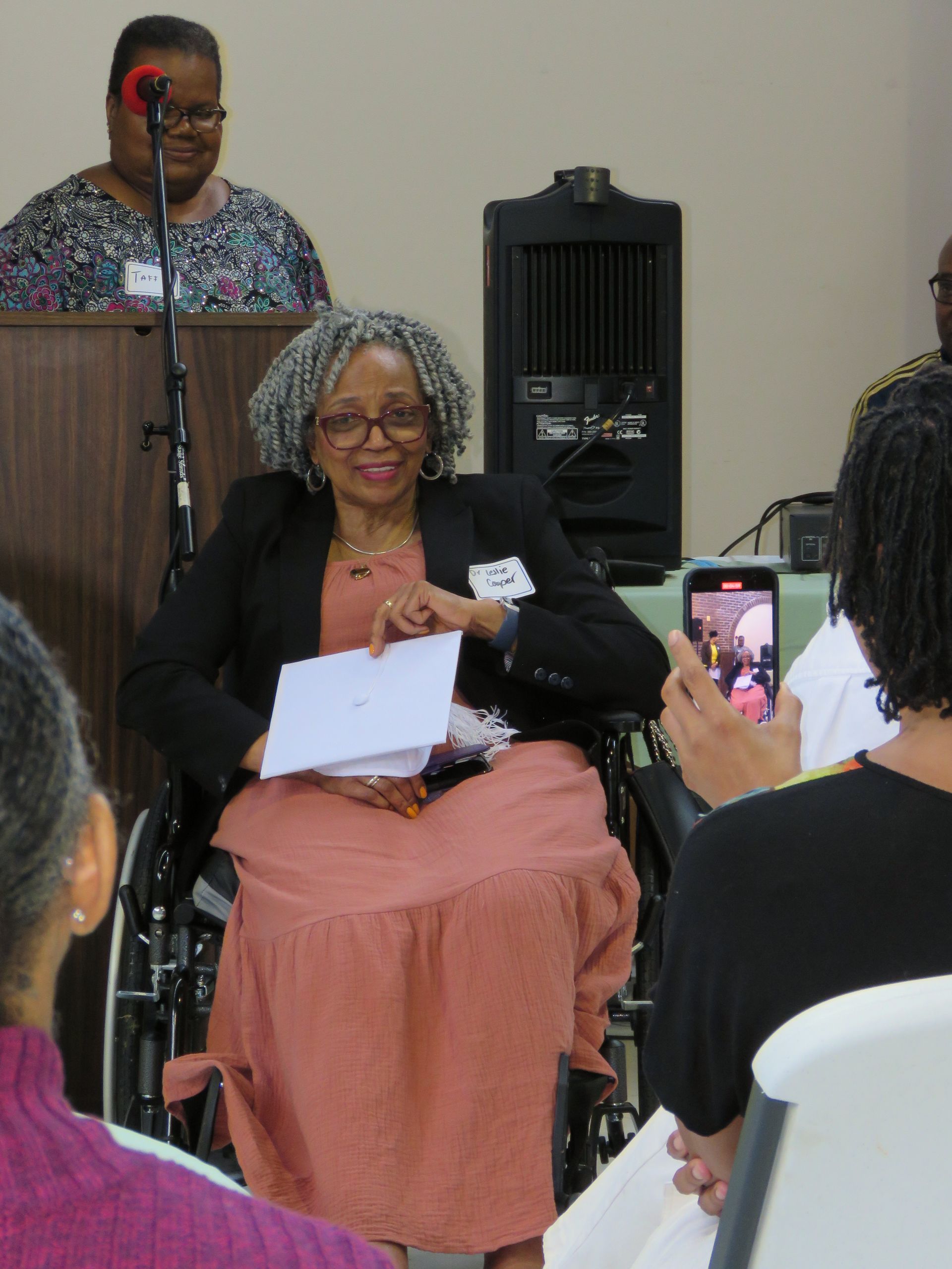 Woman in a wheelchair, holding papers, smiles. Another woman speaks at a podium. Someone films with a phone.