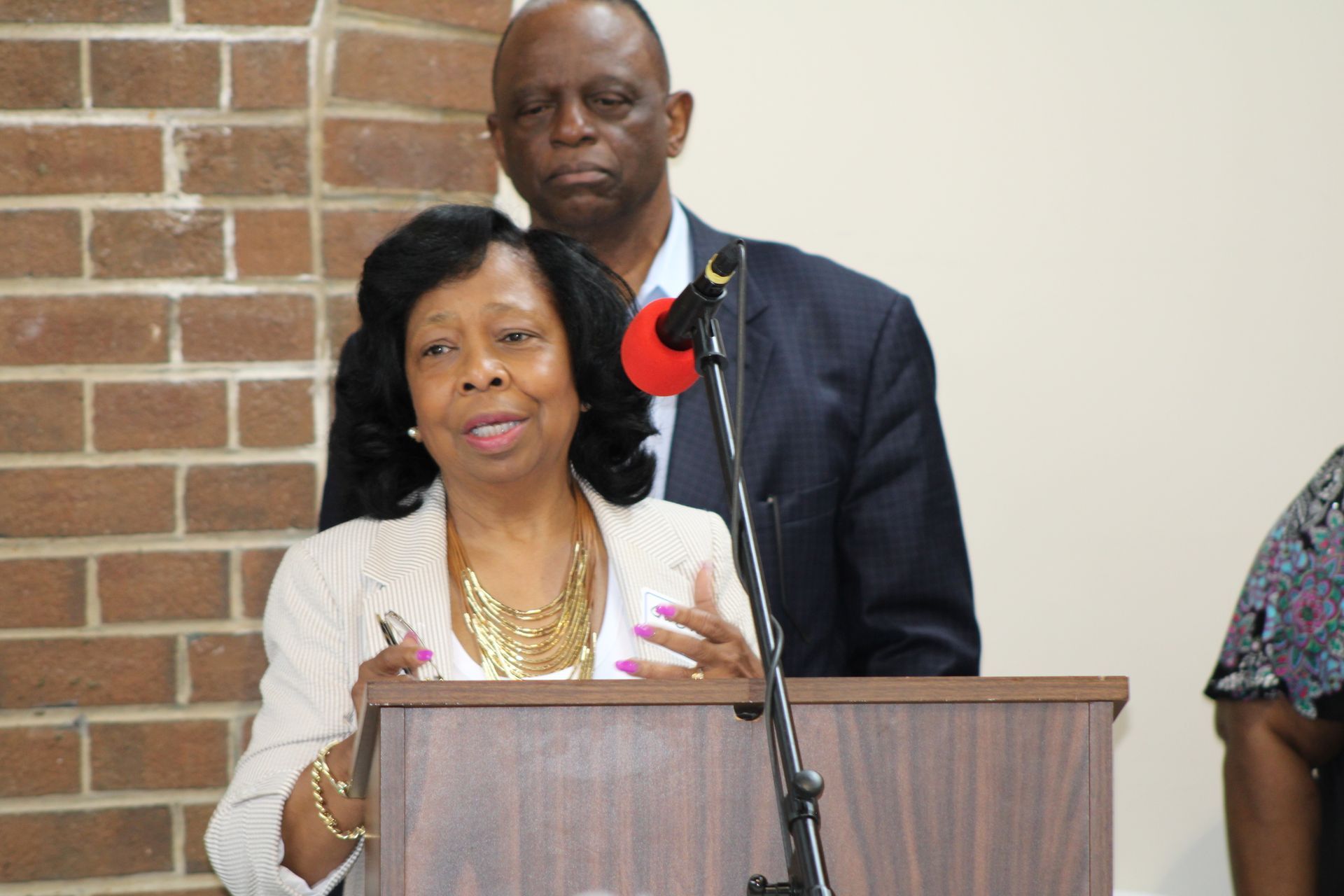 Woman speaking at a podium, man behind her. Brick wall in the background.