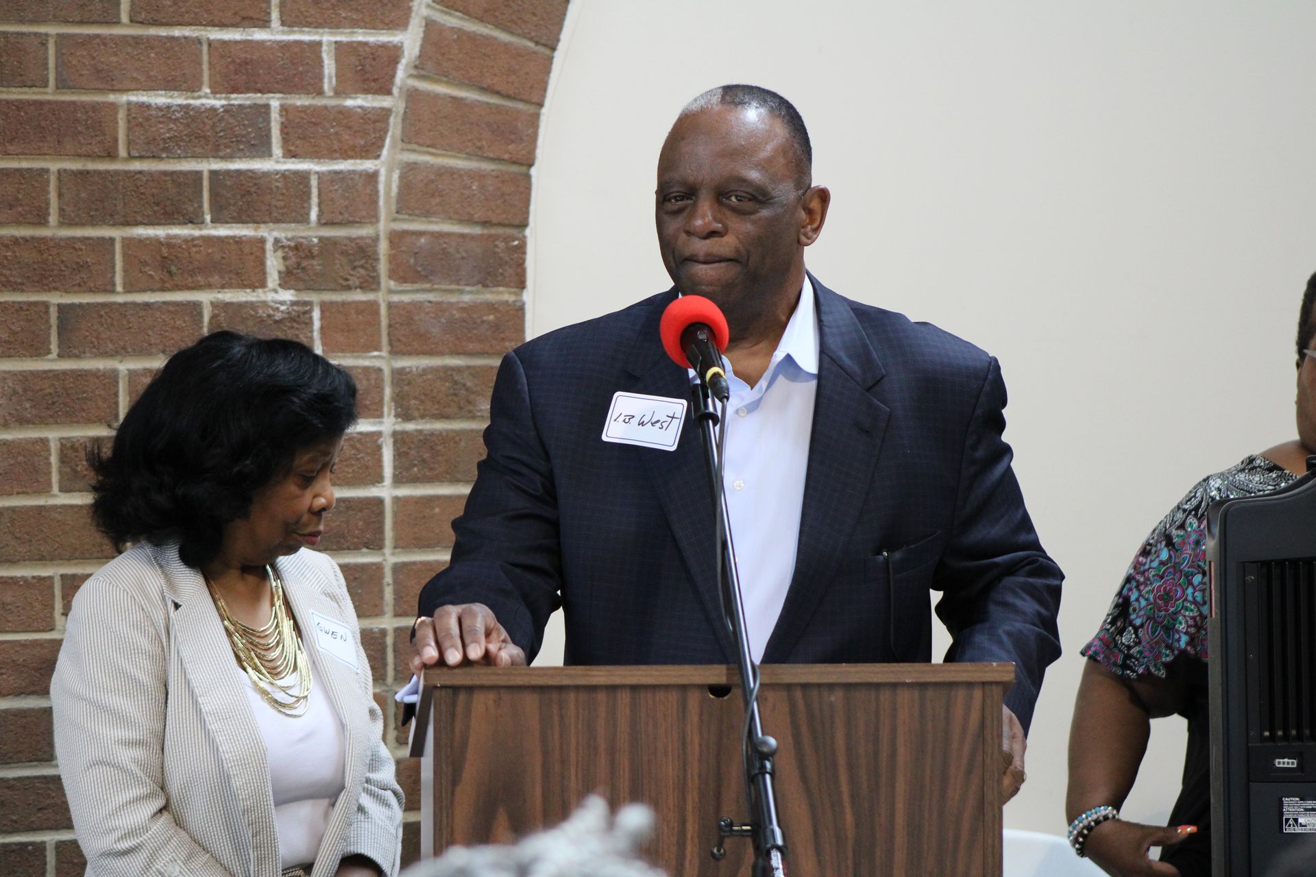 Man speaking at a podium with two people beside him, brick wall background.