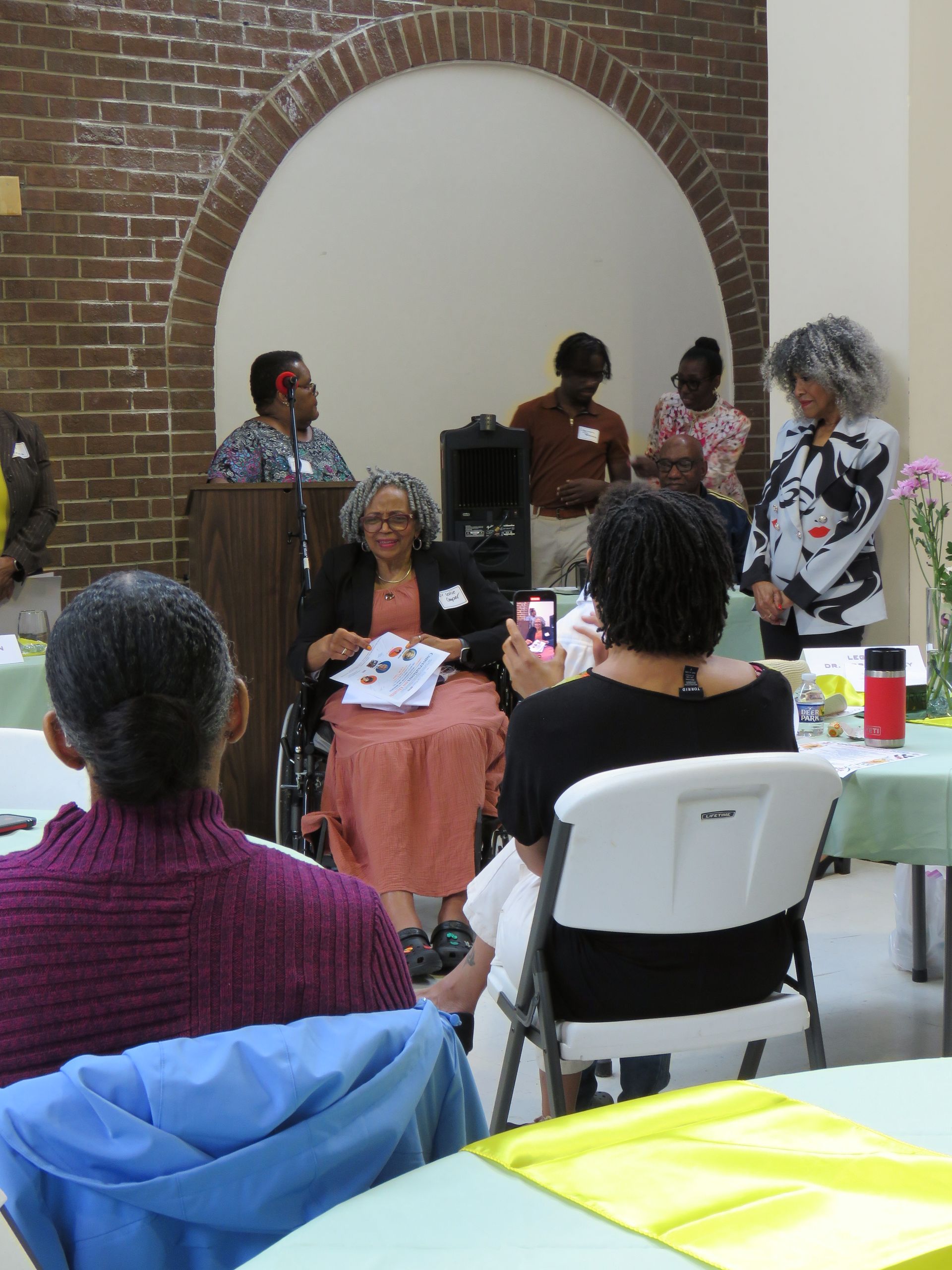 People at a gathering with a speaker in a wheelchair, under an arched brick structure.